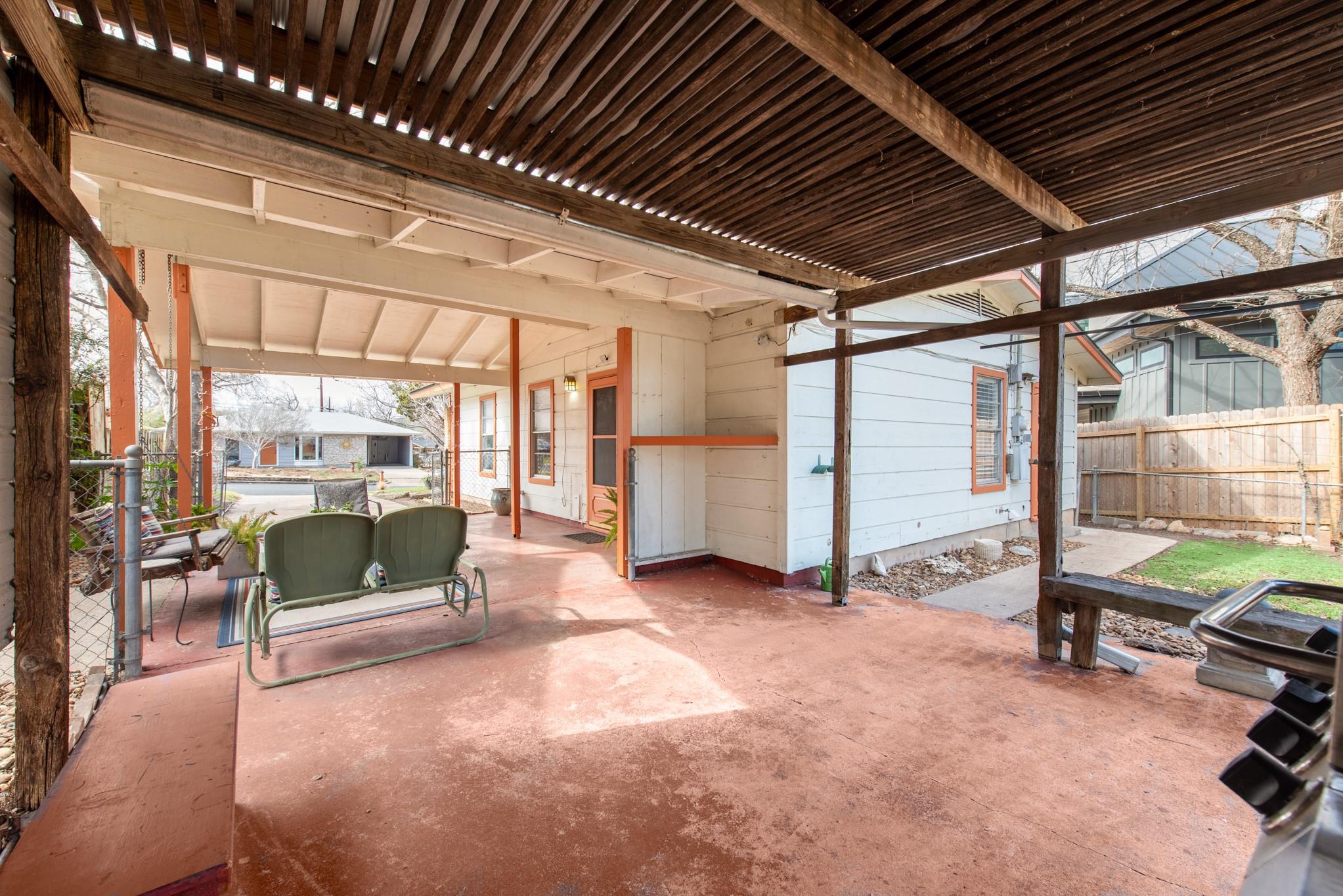 Covered patio with a dark slatted pergola roof, red concrete floor, and green metal glider.