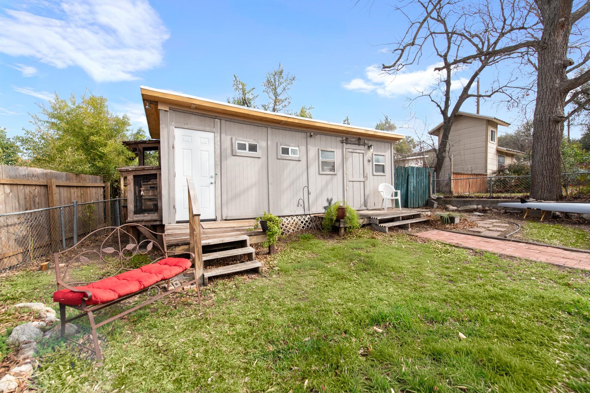 Home’s rear exterior with a raised wooden deck, grassy yard, red-cushioned bench, and large tree.