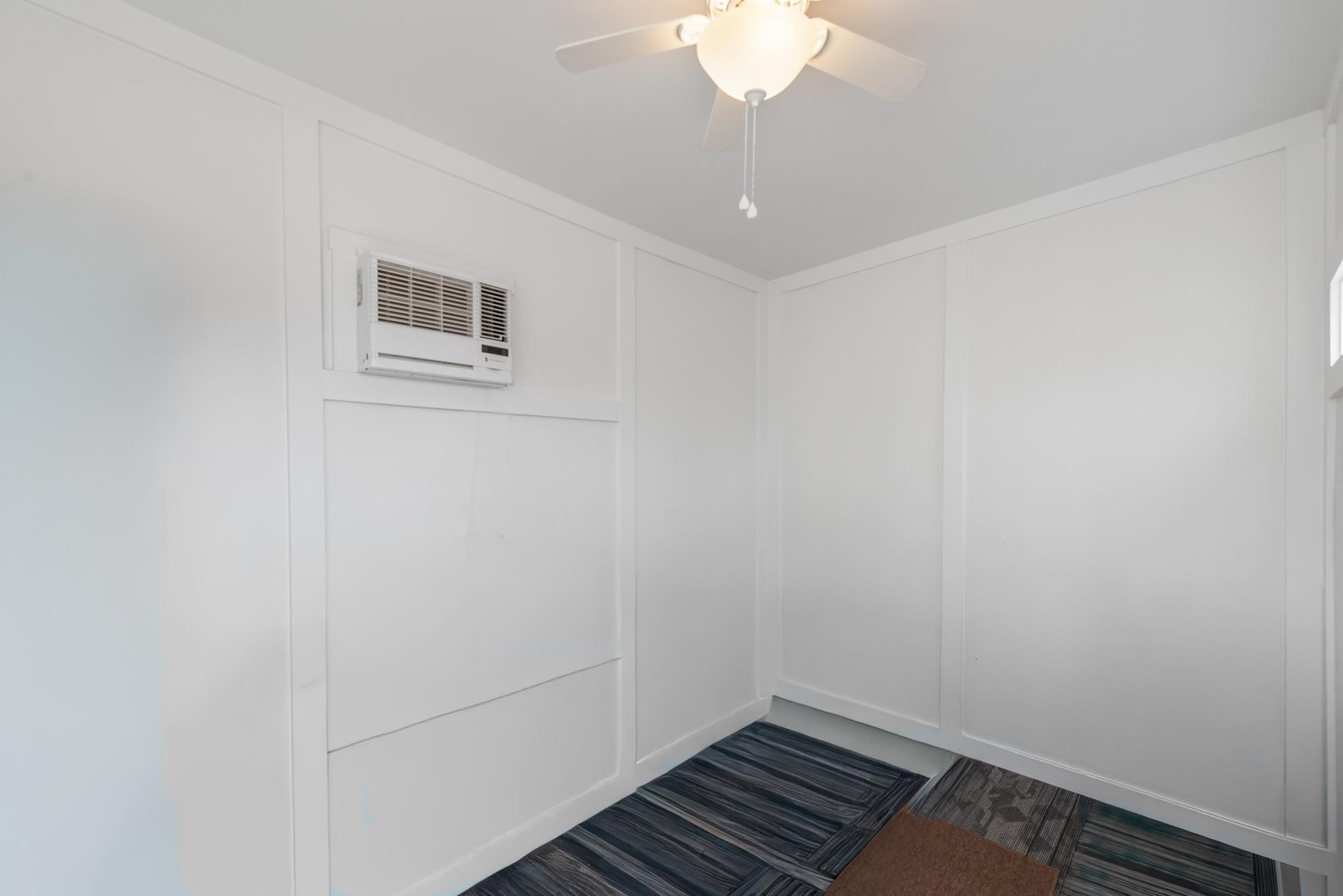 Empty white room with wall paneling, a window air conditioner, ceiling fan, and dark patterned carpet tiles.