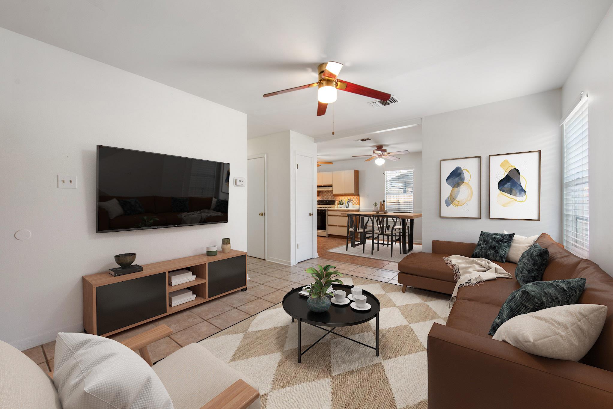 Staged living room with a brown sectional and television, opening to a dining area and kitchen.