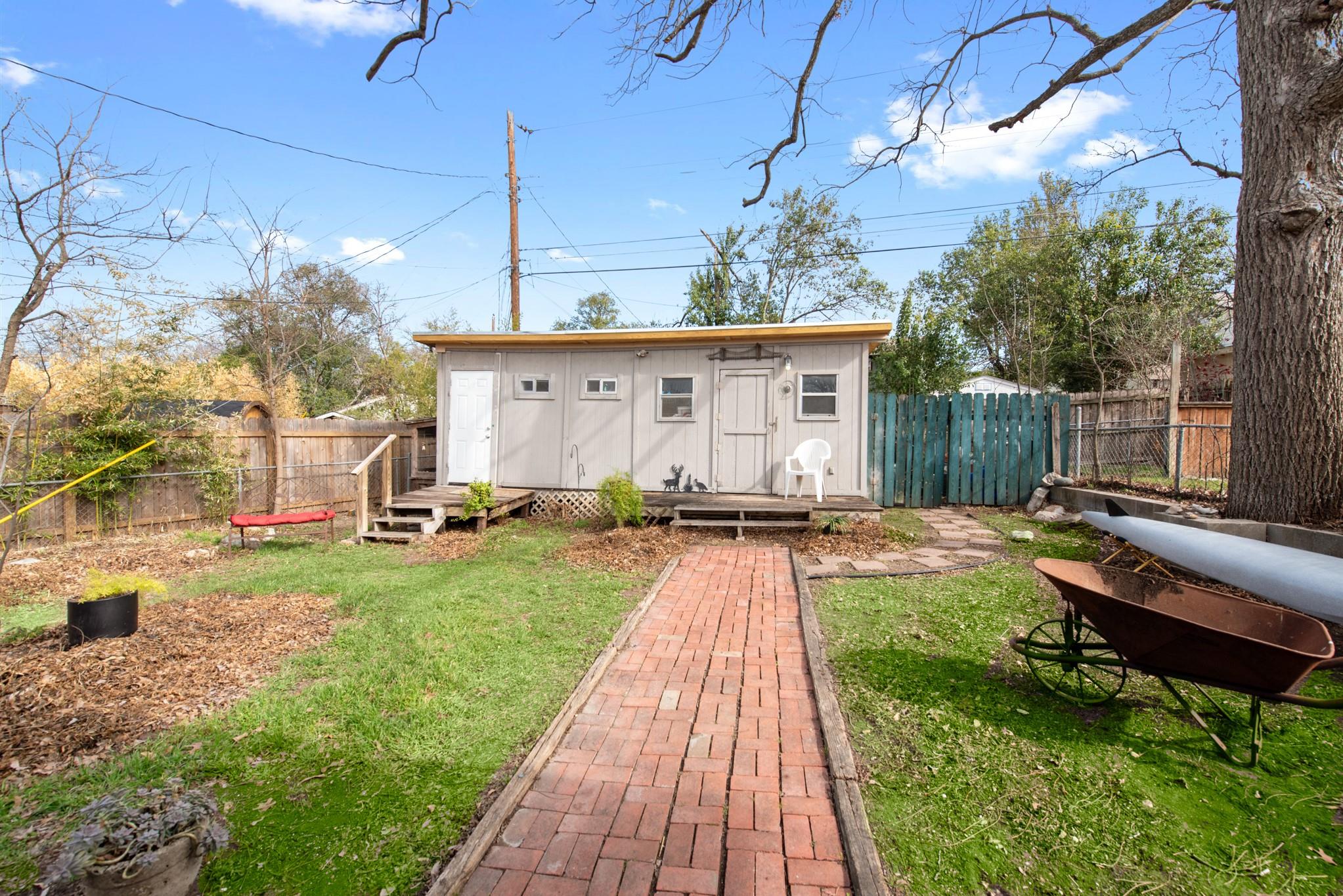 Gray backyard outbuilding with a low deck, reached by a brick walkway through the lawn.