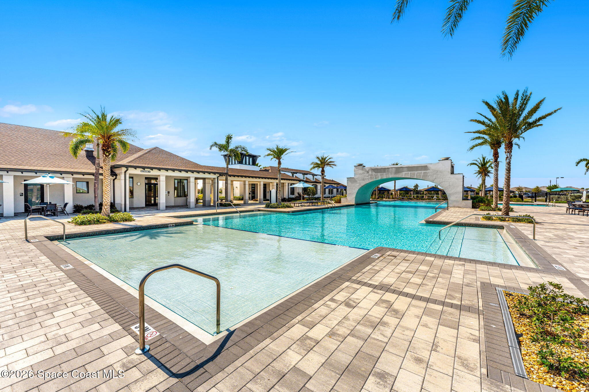 Community pool with a shallow entry, stone arch, palm trees, and spacious paved deck.