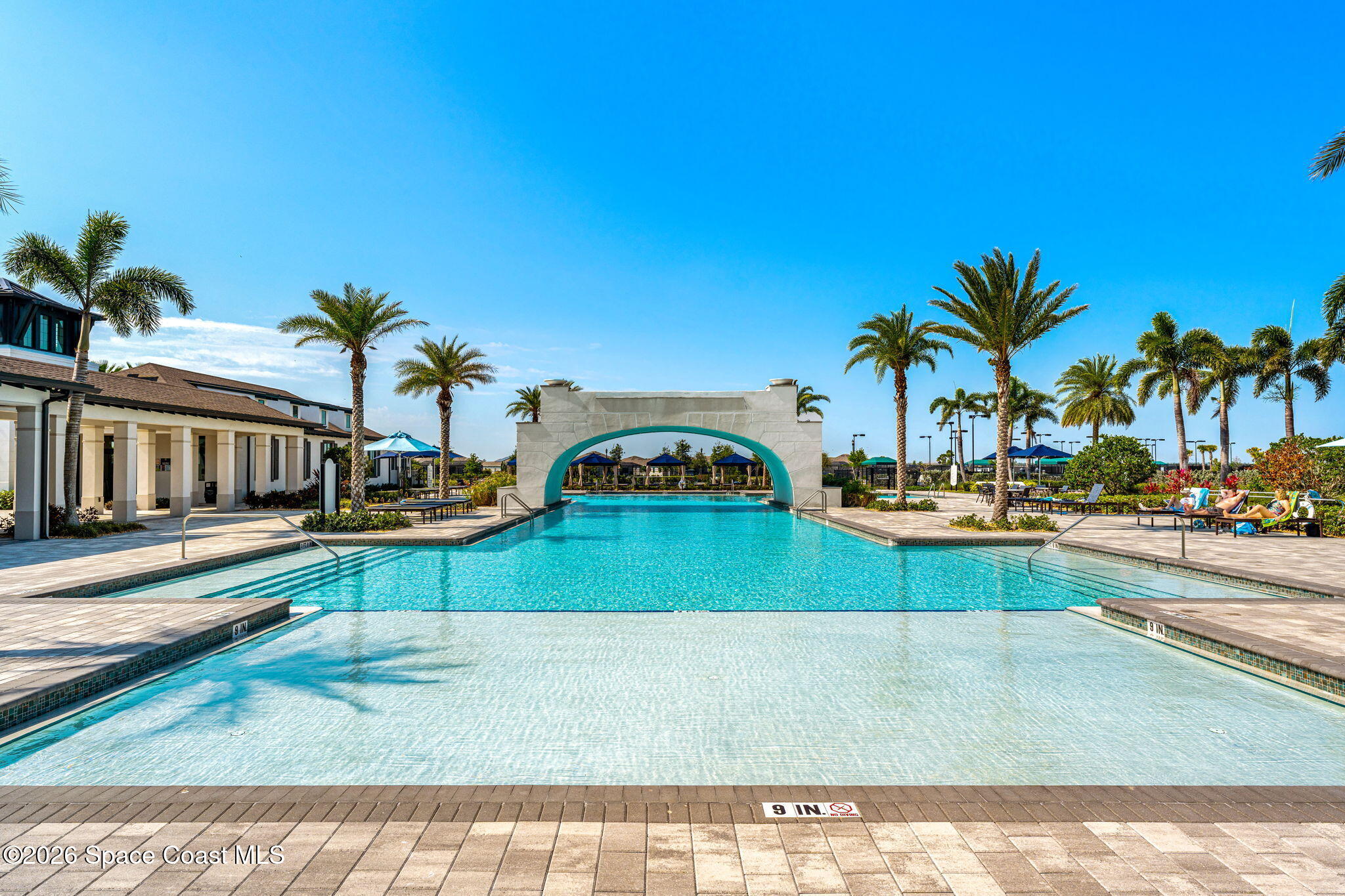 Outdoor pool with a shallow-entry area, turquoise arch, palm trees, and clubhouse.