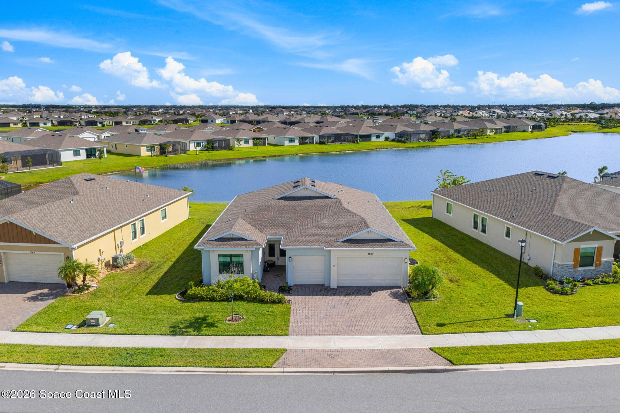 Aerial view of a single-story home beside a lake, with a paved driveway and neighboring homes beyond.