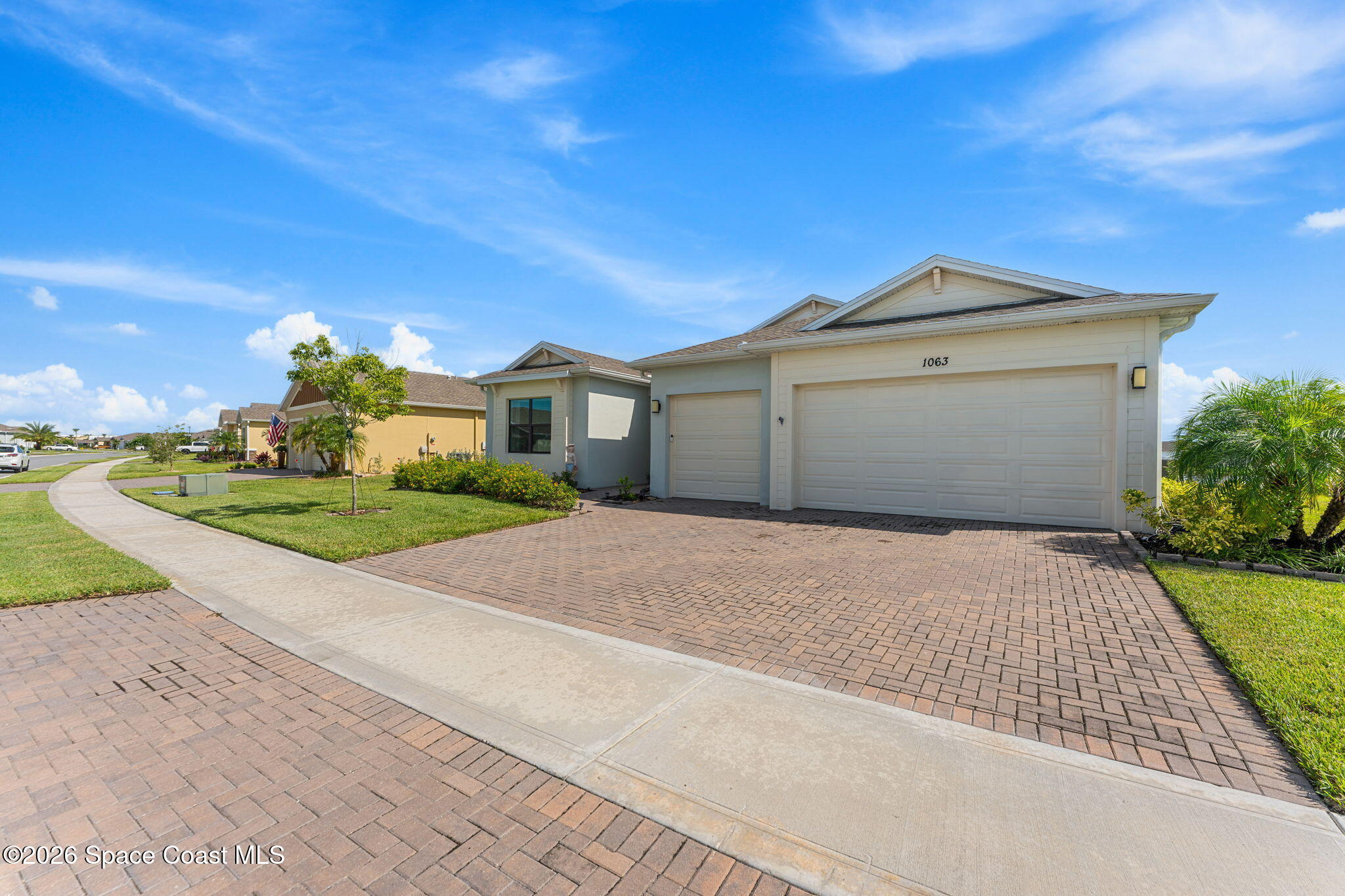 Home exterior with a three-car garage, brick-paver driveway, and tropical landscaping.