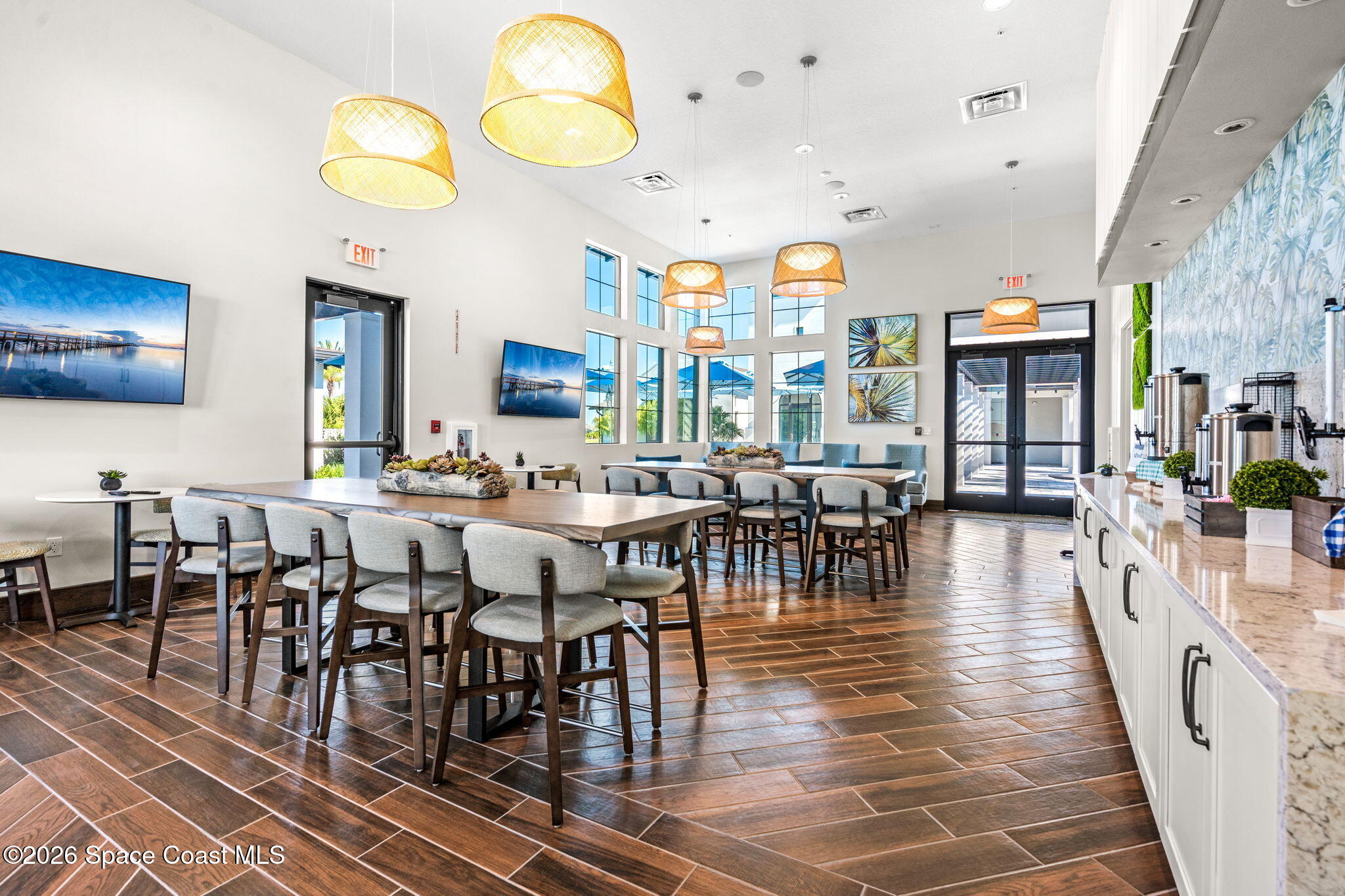 Furnished communal dining area with long tables, tall windows, pendant lights, and a coffee service counter.