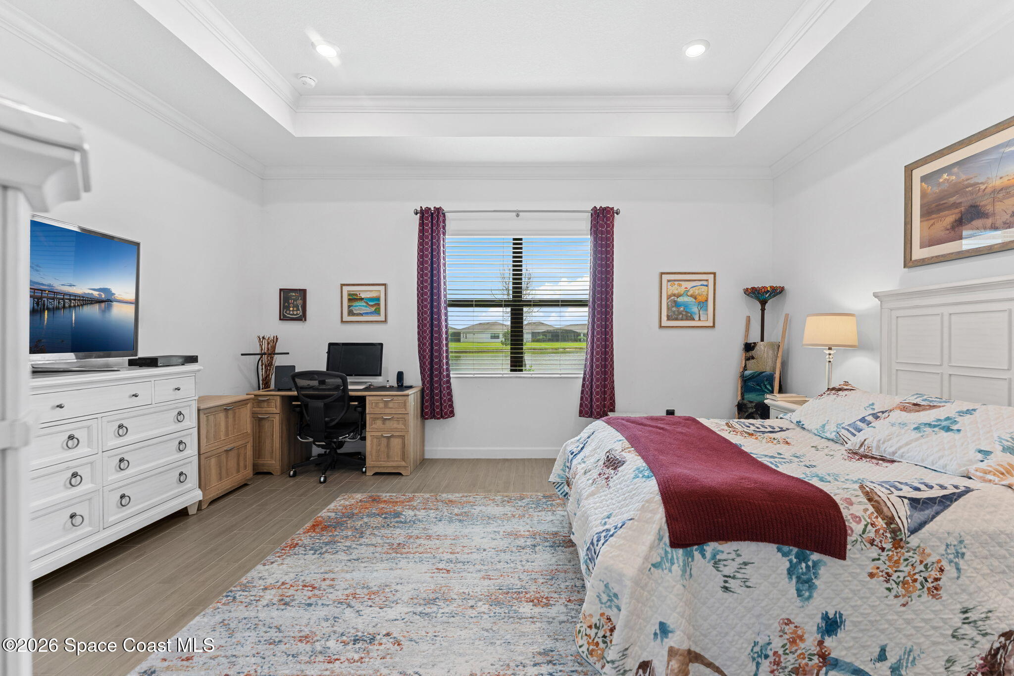 Furnished bedroom with a floral quilted bed, burgundy runner, desk, and large window.