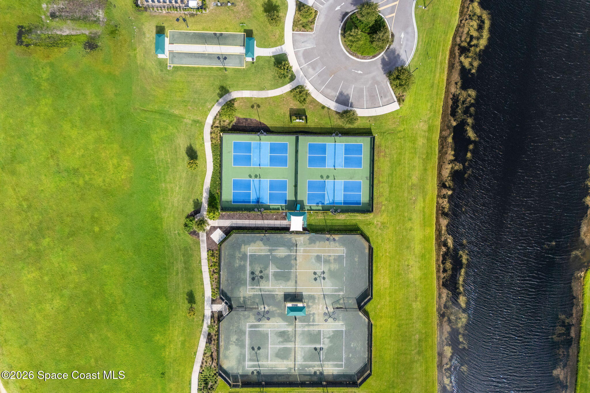 Aerial view of community courts beside a body of water, with walking paths and a circular parking area.