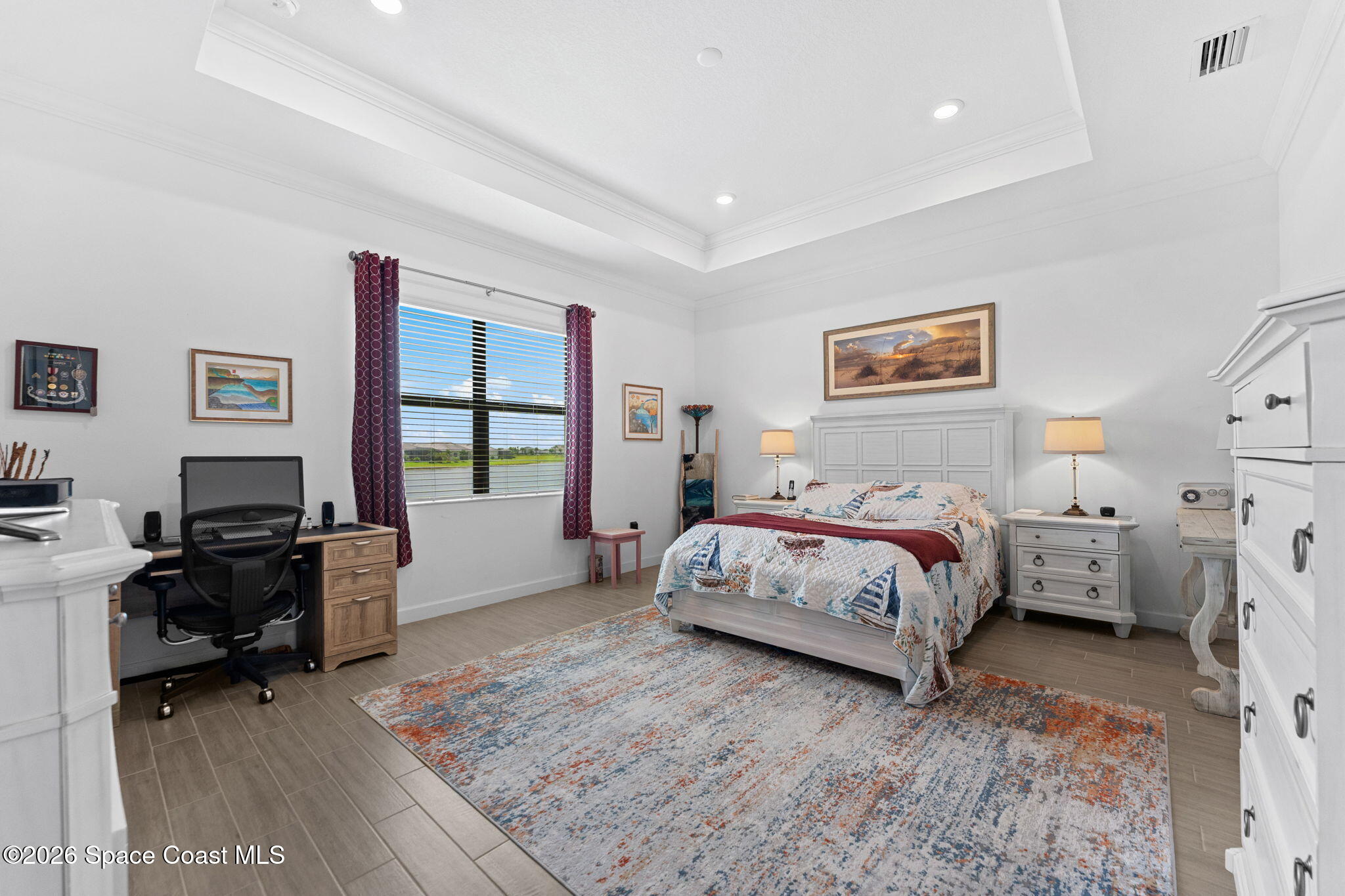 Furnished bedroom with a tray ceiling, desk, patterned rug, and window overlooking water.