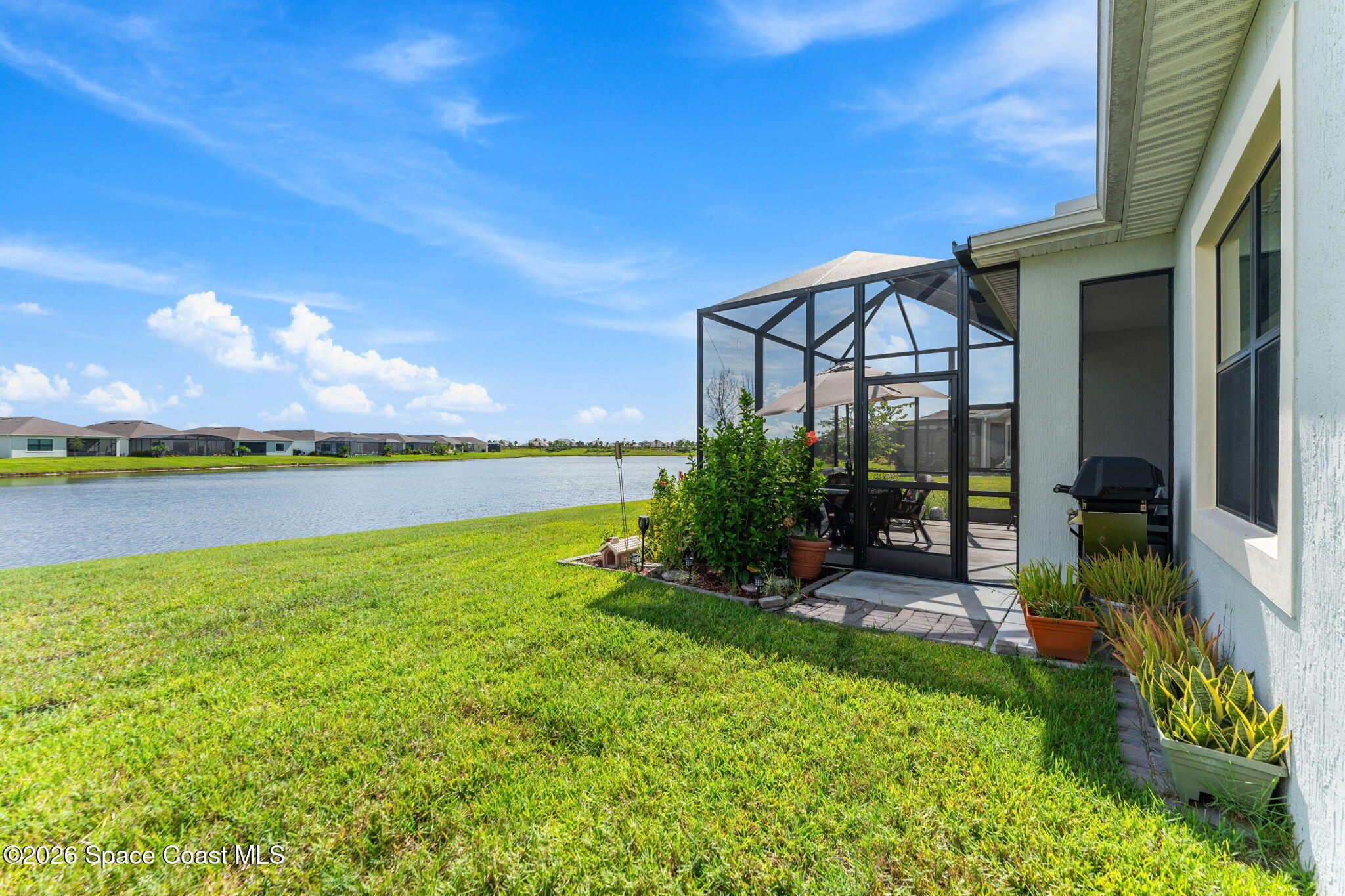 Home’s screened patio and lawn overlook a lake, with outdoor seating, potted plants, and a grill.
