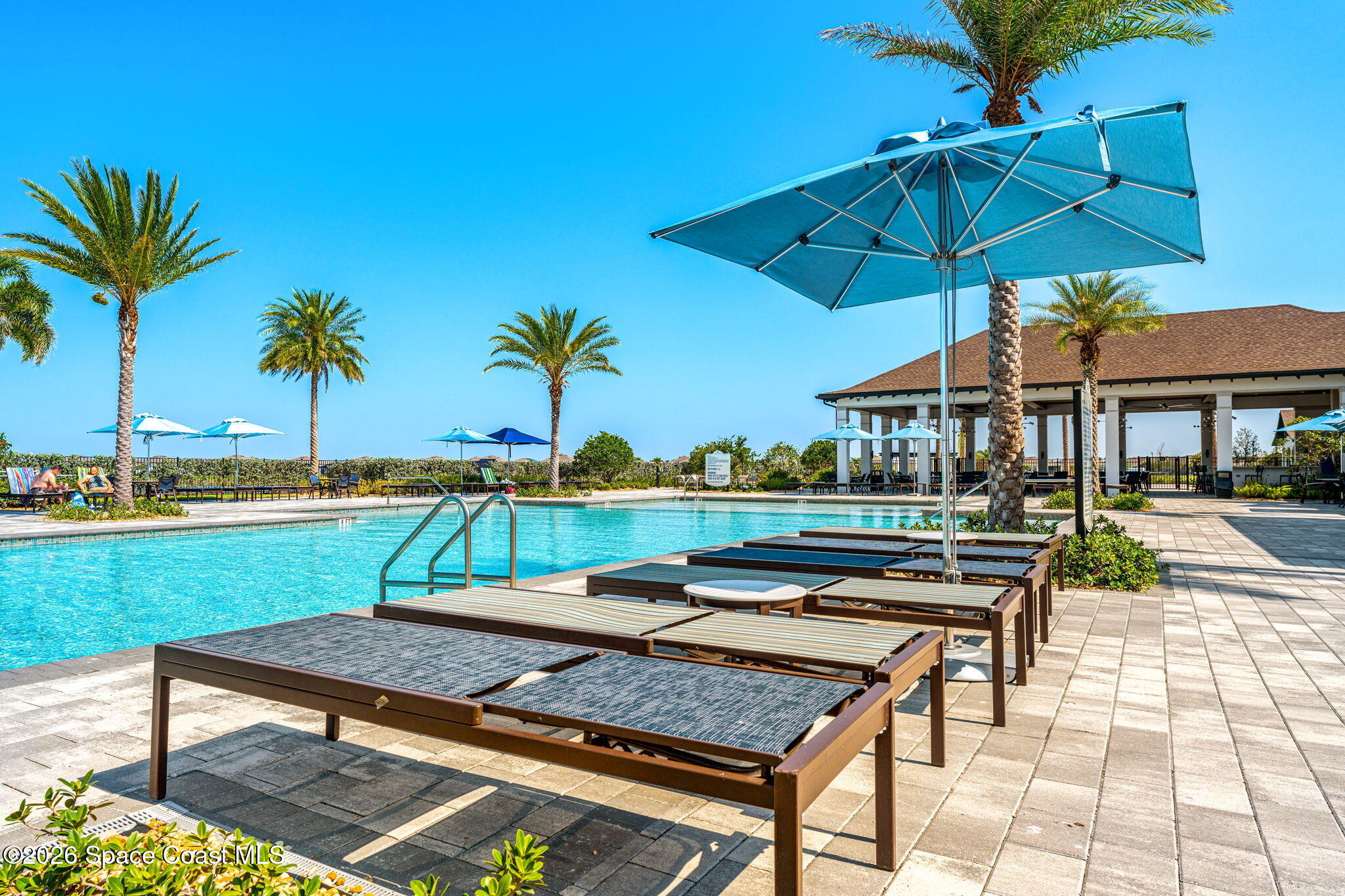 Community pool with blue umbrellas, palm trees, lounge chairs, and a covered pavilion.