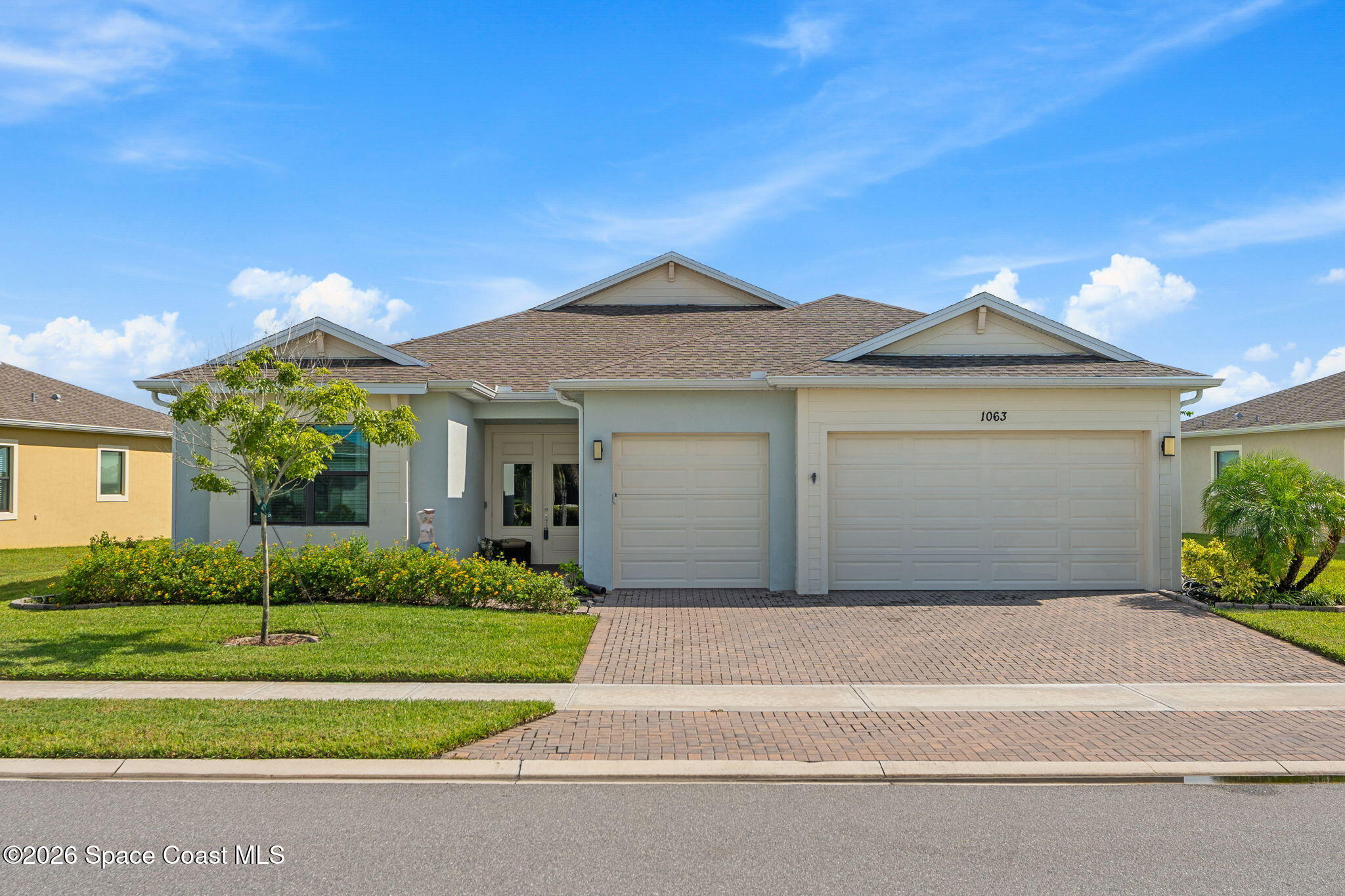 Single-story gray house with two garage doors, a brick-paved driveway, and a landscaped front lawn.