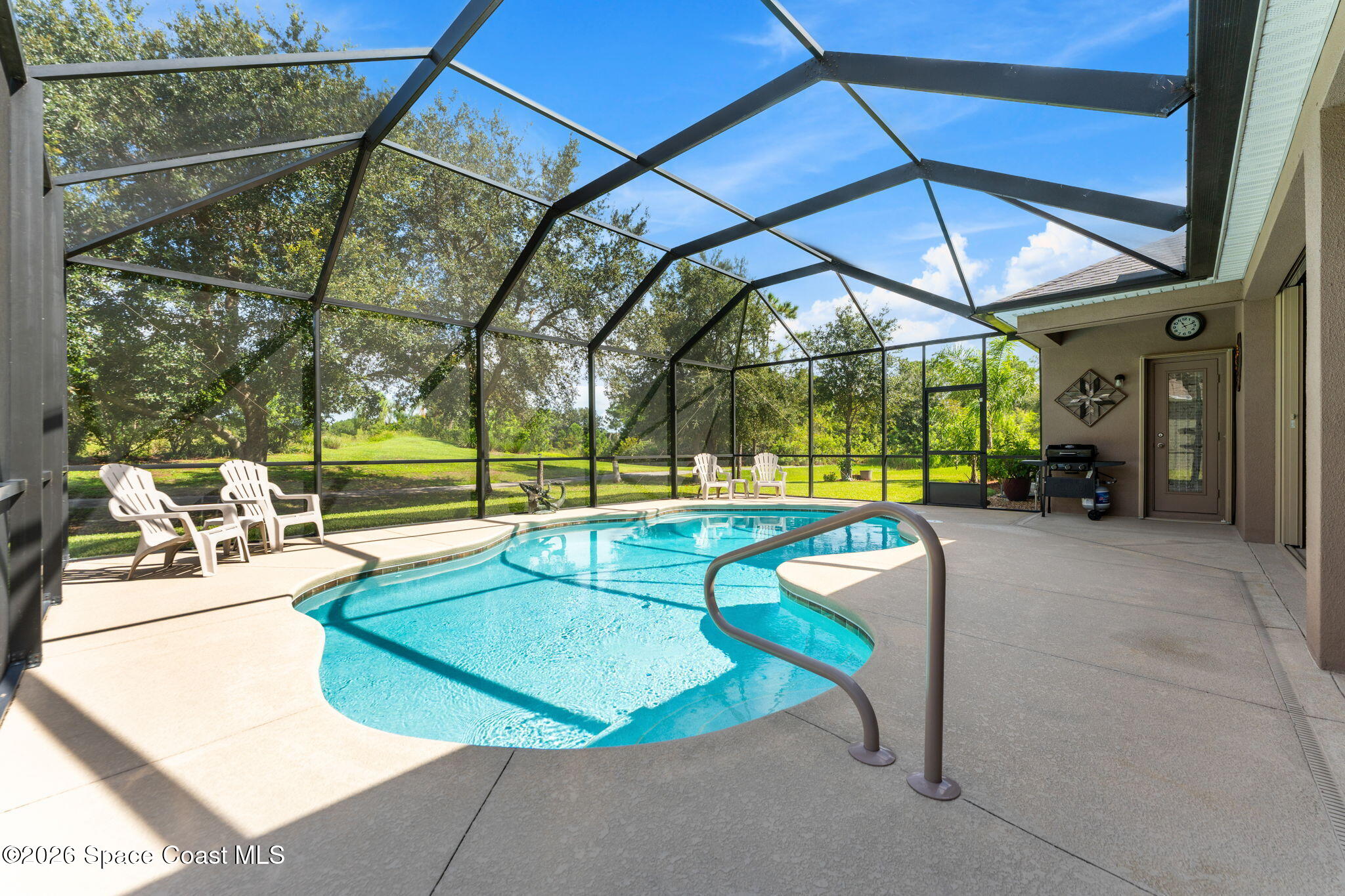 Screened-in swimming pool with a curved shape, patio chairs, and a sunny concrete deck.