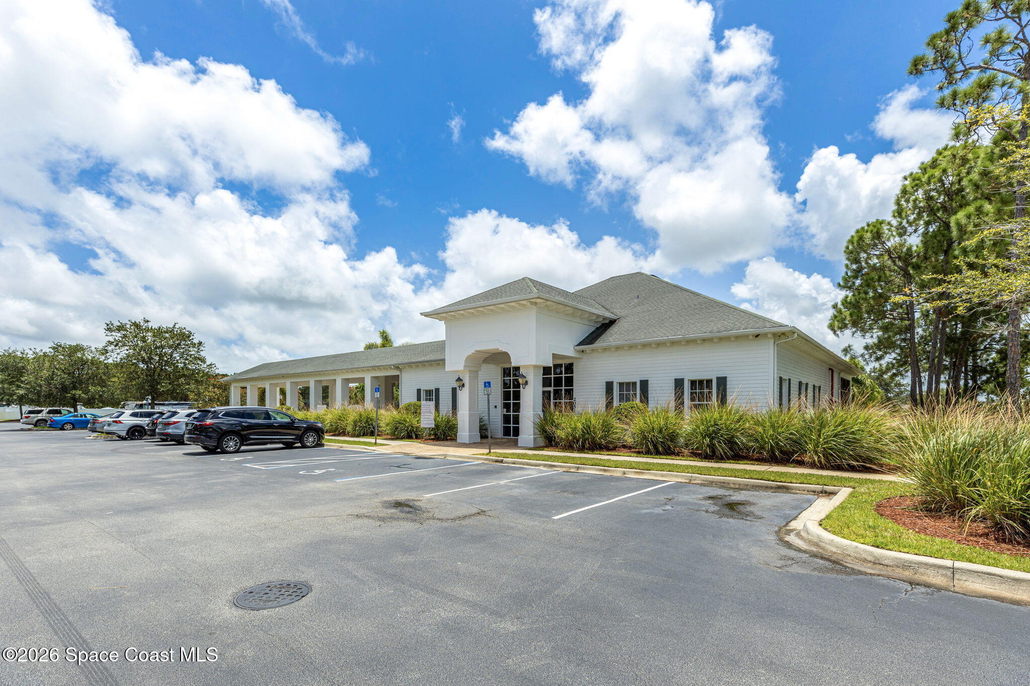 White building with a covered entrance, landscaped grasses, and parking spaces in front.