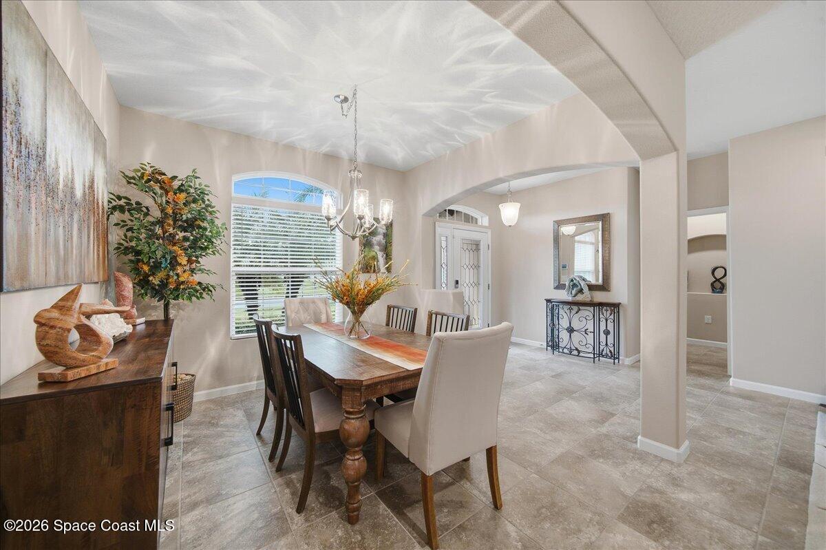Dining room with a long wooden table, mixed chairs, tiled floors, and an arched opening to the entry hall.