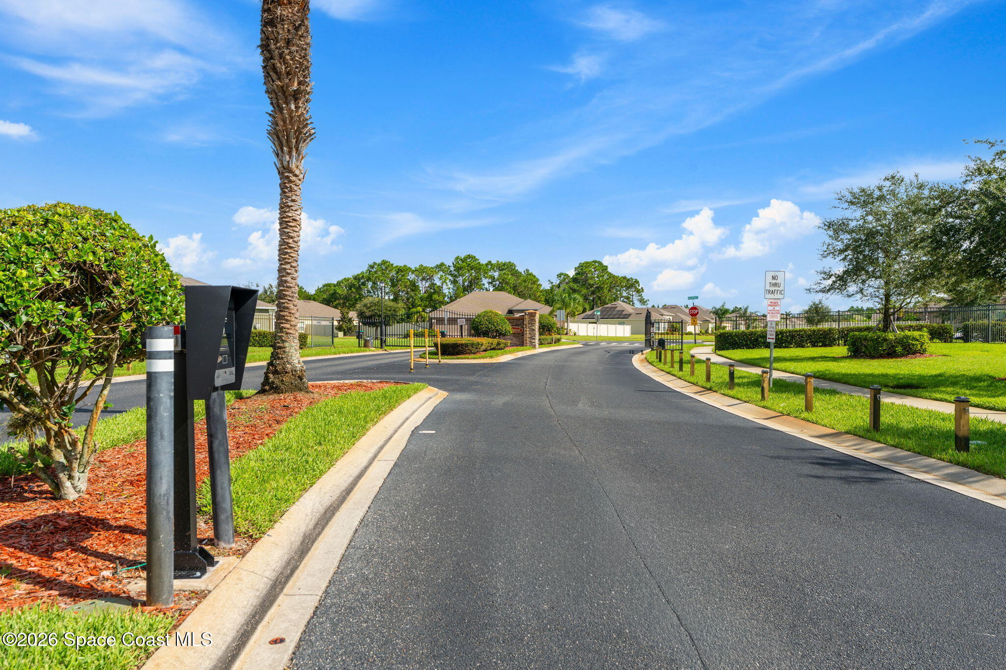 Residential community entrance road with a gate, palm tree, and no-through-traffic sign.