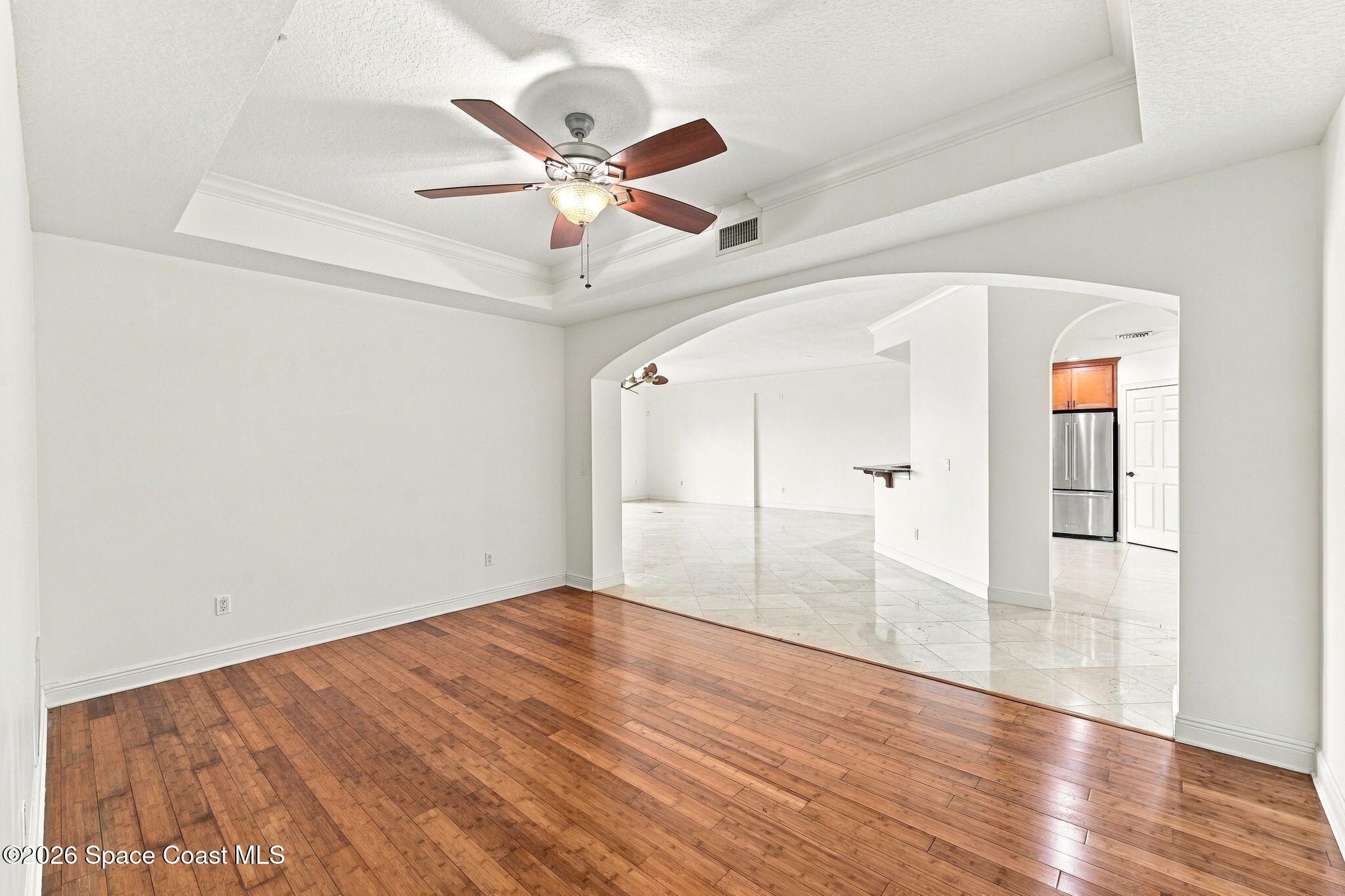 Empty living room with hardwood floors, tray ceiling, and arched opening to a tiled kitchen.