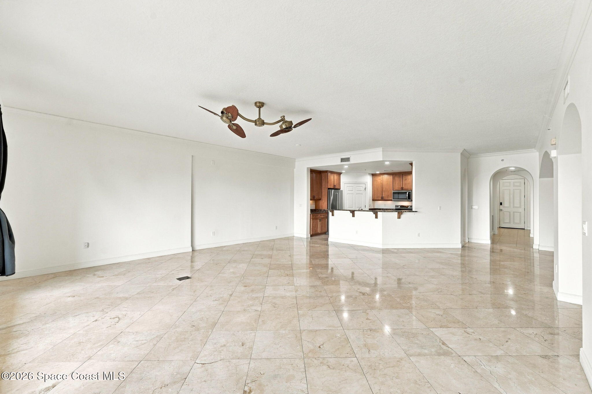 Empty living area with beige tile floors, a ceiling fan, and an open kitchen with wood cabinets.