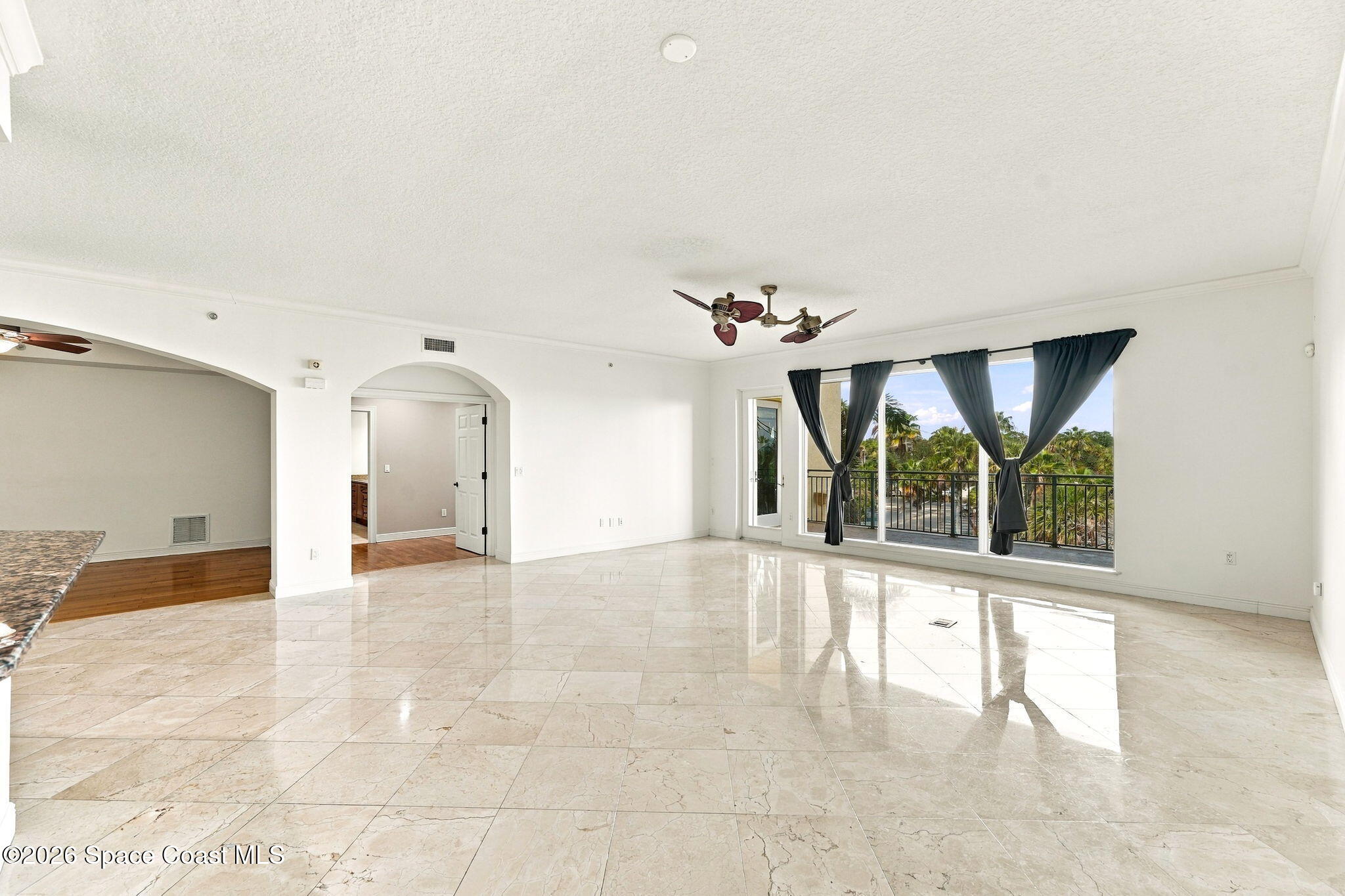 Vacant living room with polished marble tile floors and curtained doors opening to a palm-lined balcony.