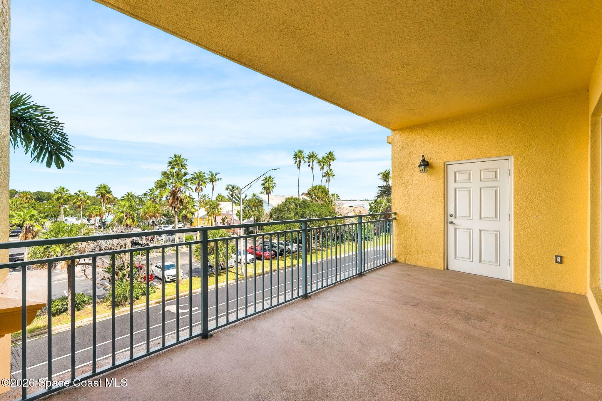 Covered balcony with a black railing overlooking palm trees, rooftops, and a parking area.