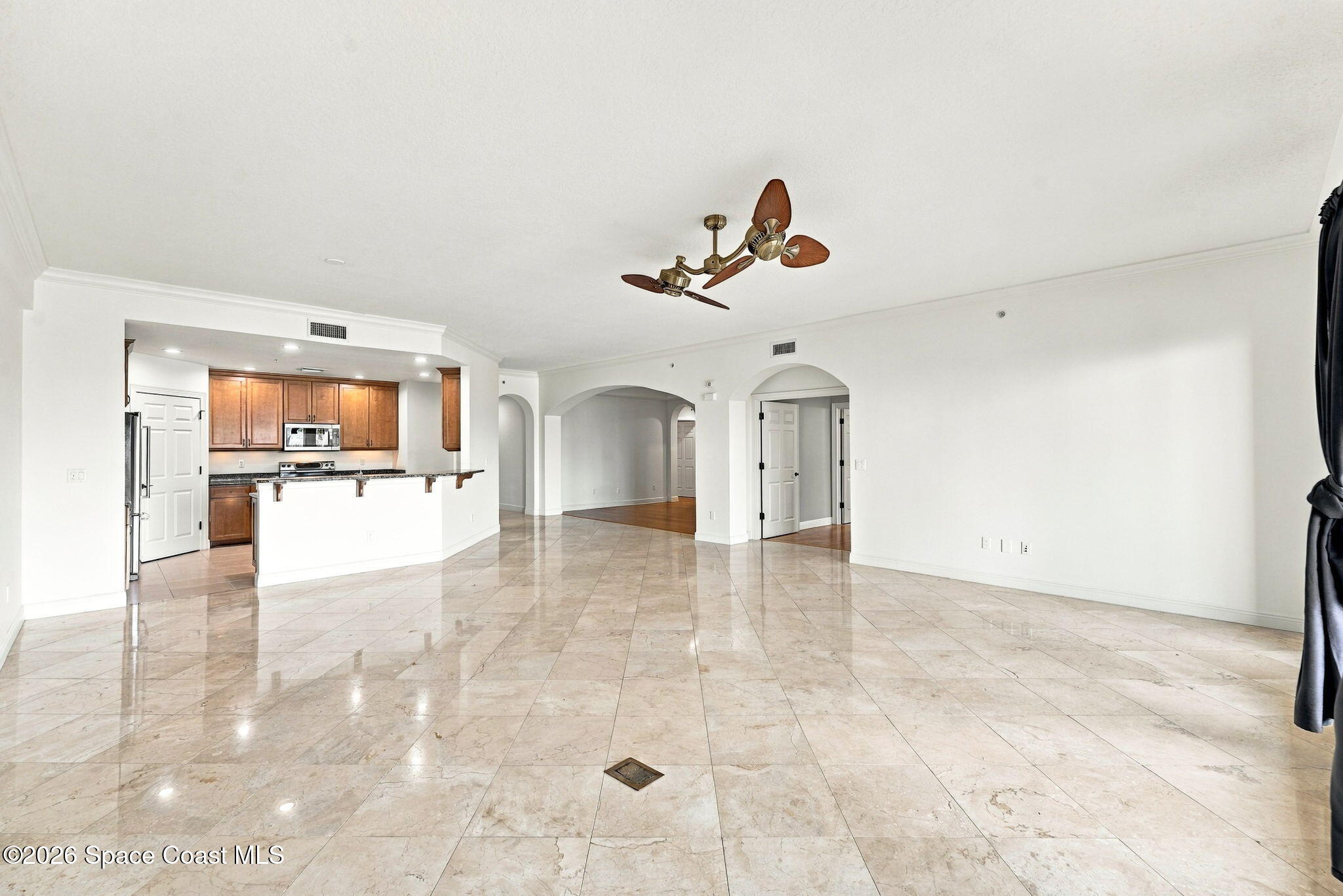 Empty open-plan living area with glossy stone tile floors, arched doorways, and an adjoining wood-cabinet kitchen.