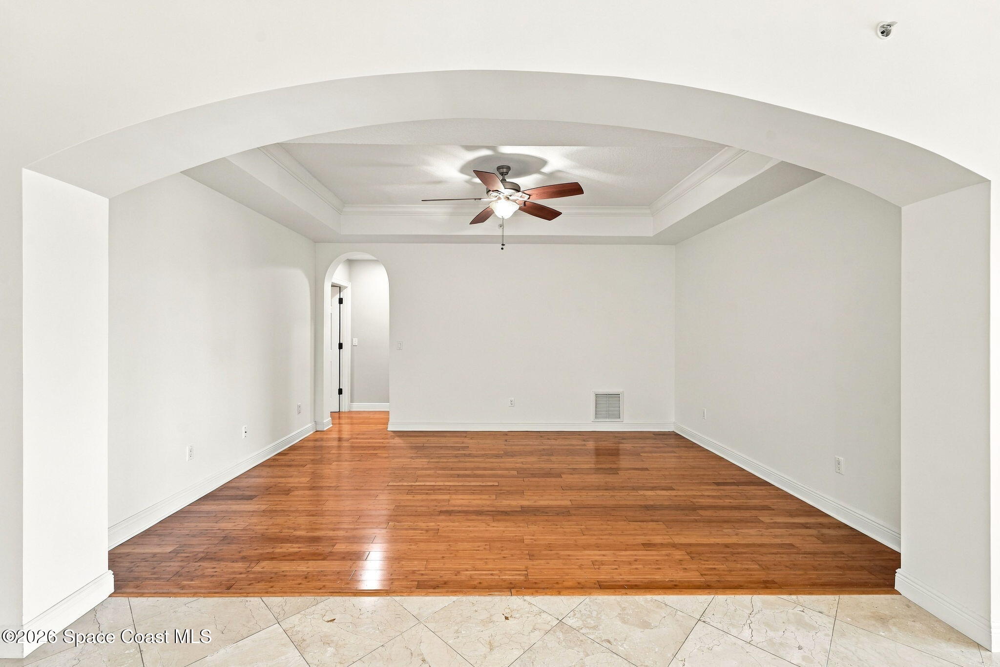 Vacant living room with hardwood floors, a tray ceiling, ceiling fan, and arched doorway.
