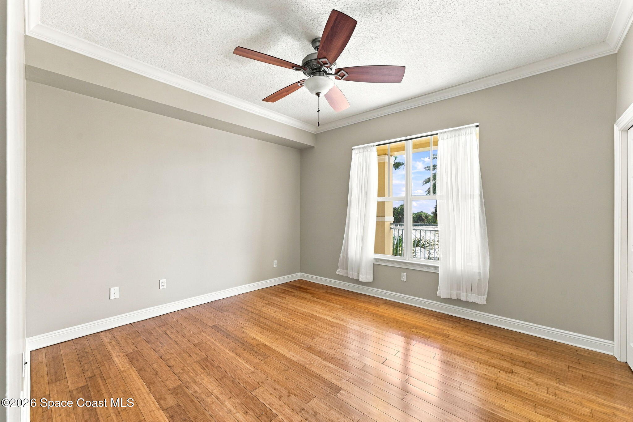 Empty room with warm wood floors, a ceiling fan, and a curtained window overlooking a balcony.