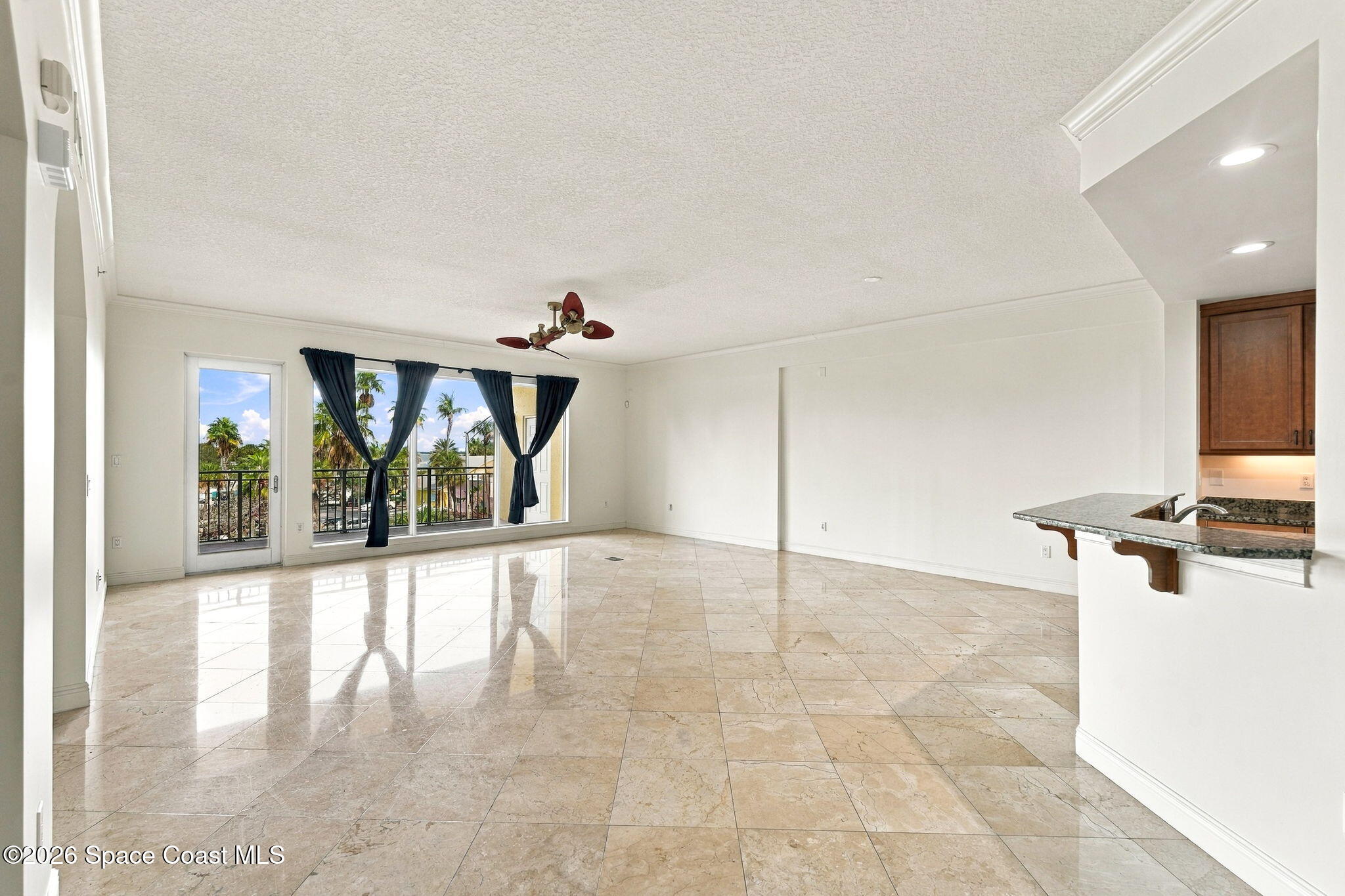 Empty living area with glossy tile floors, balcony doors, palm trees outside, and a granite breakfast bar.
