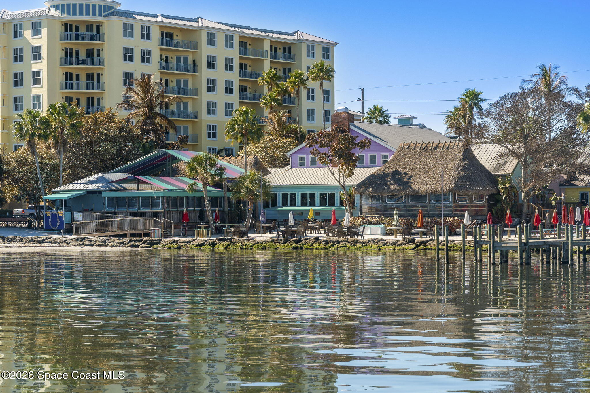 Waterfront buildings, including a yellow condominium and thatched pavilion, reflected in calm water beside a dock.