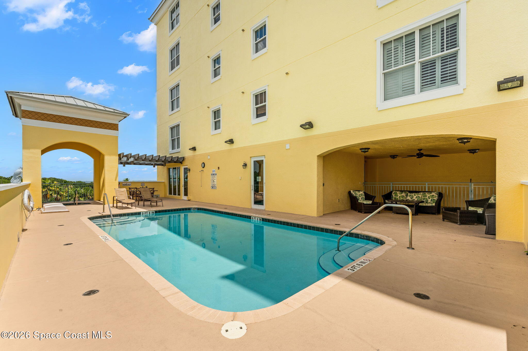 Outdoor pool beside a yellow condominium building, with a covered seating area and lounge chairs.