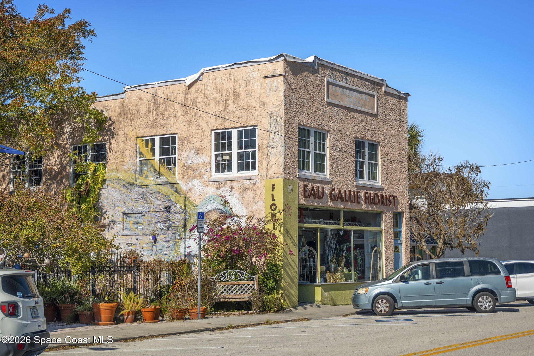 Two-story stucco building with a florist storefront, side-wall mural, and parked cars.