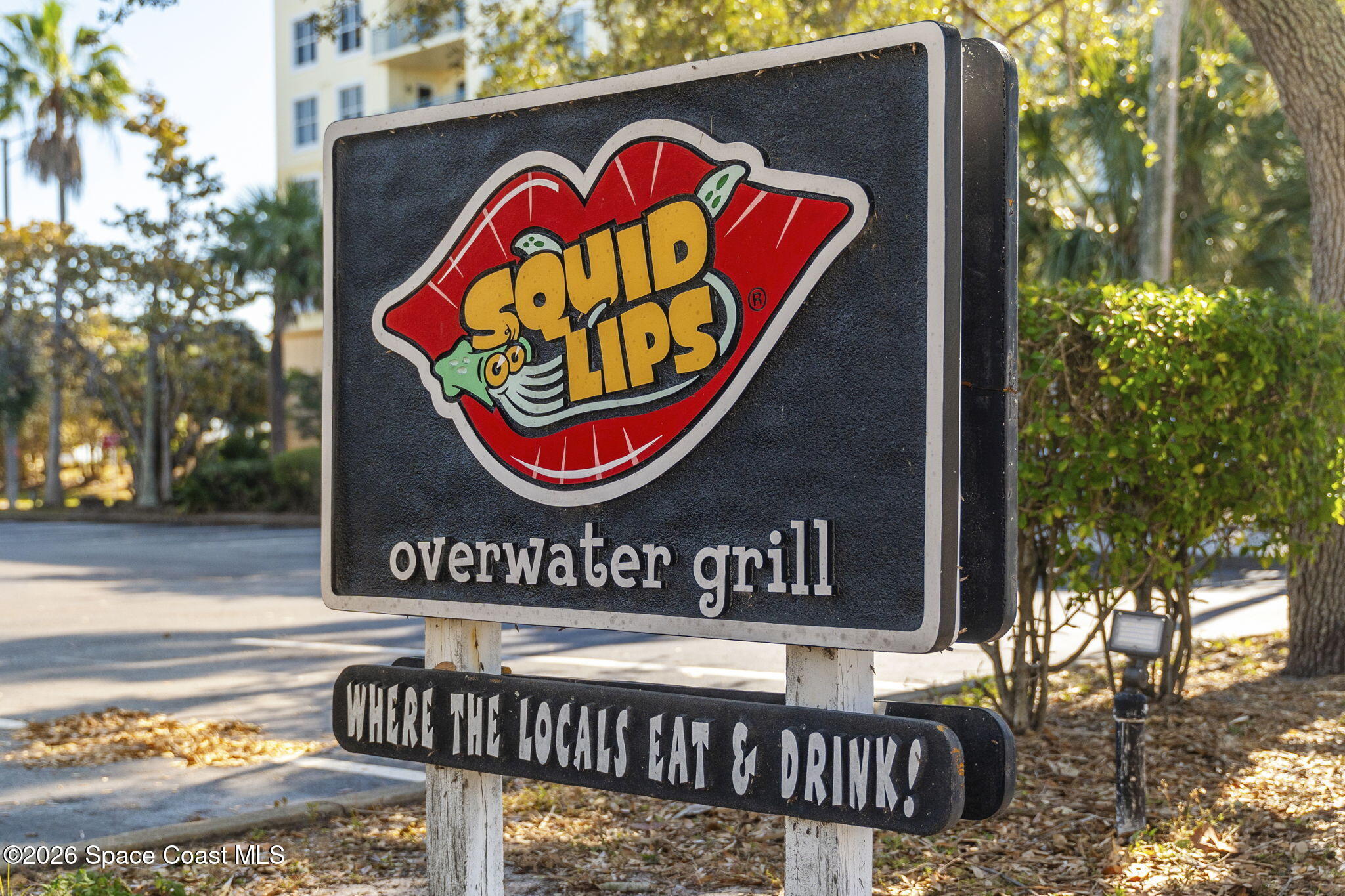 Overwater Grill sign with the Squid Lips logo beside a tree-lined road.
