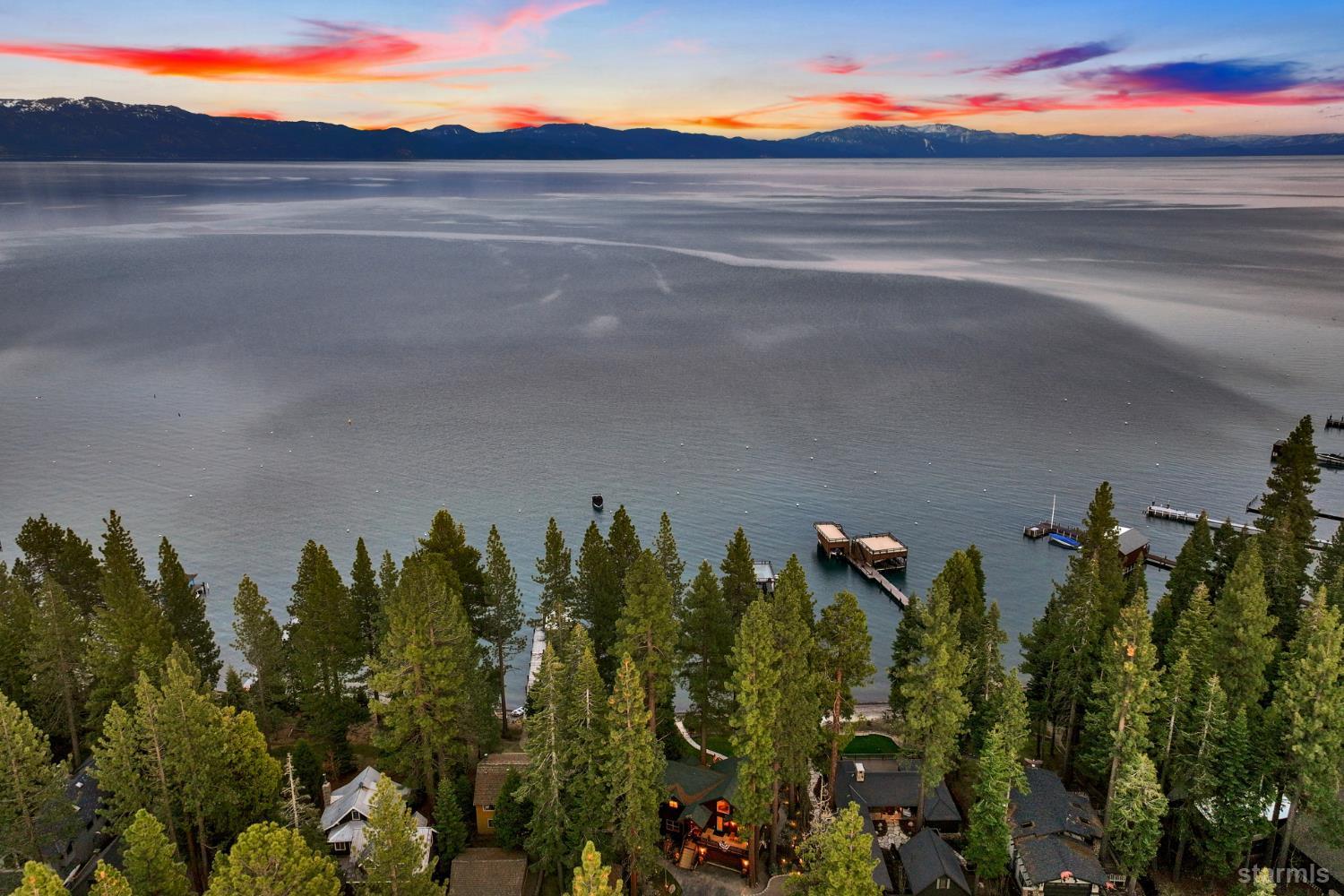 Aerial view of Lake Tahoe, with forested homes and docks below distant snow-dusted mountains at dusk.