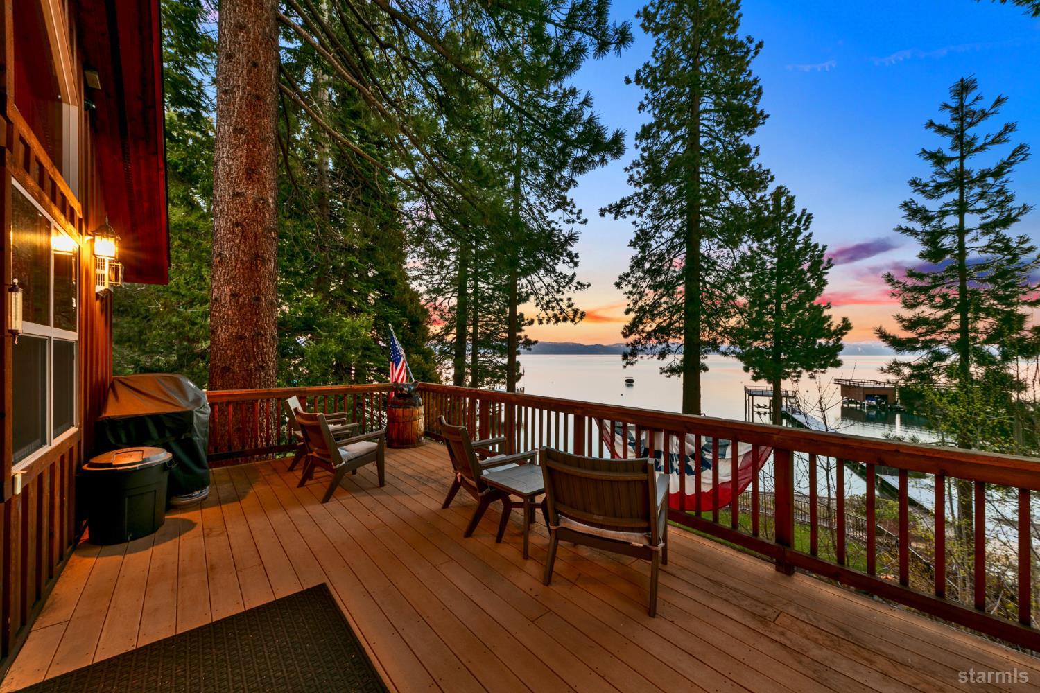 Furnished lakeside deck with Adirondack chairs and pine trees overlooking the water at sunset.