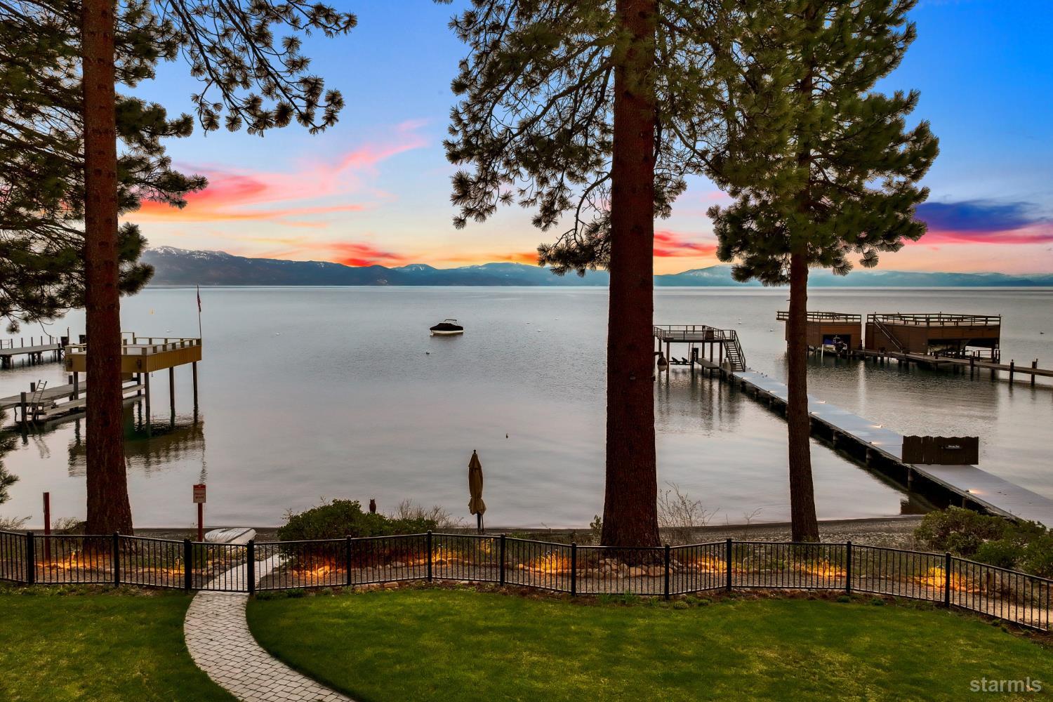 Lakefront yard with docks, pine trees, and distant mountains beneath a colorful dusk sky.