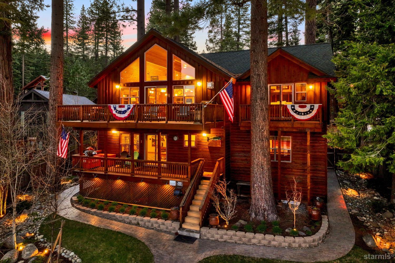 Wood cabin with expansive balconies, American flags, and warm lights among pine trees at dusk.