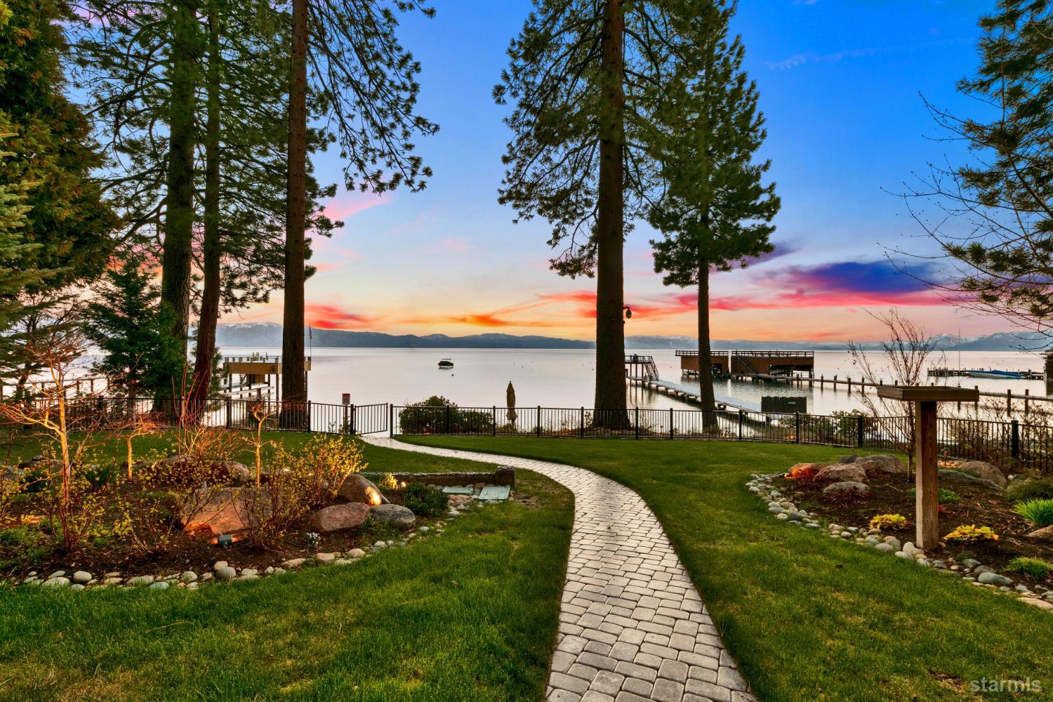Lakeside yard with a curved stone path, pine trees, and docks on the water at sunset.