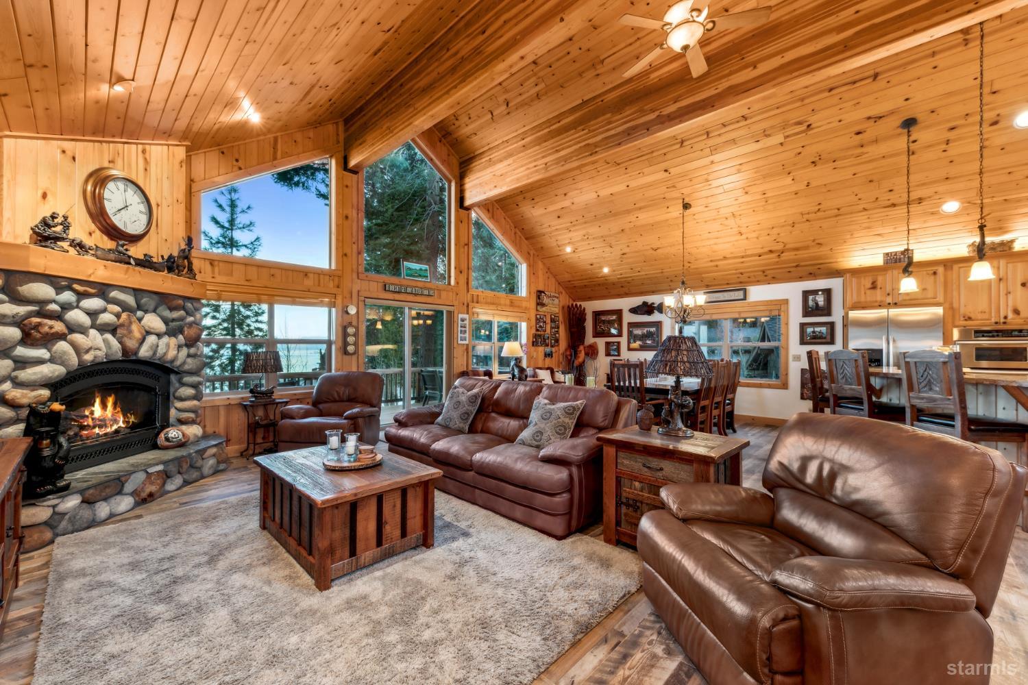 Open-plan living room with vaulted pine ceiling, stone fireplace, leather seating, and windows overlooking trees and water.