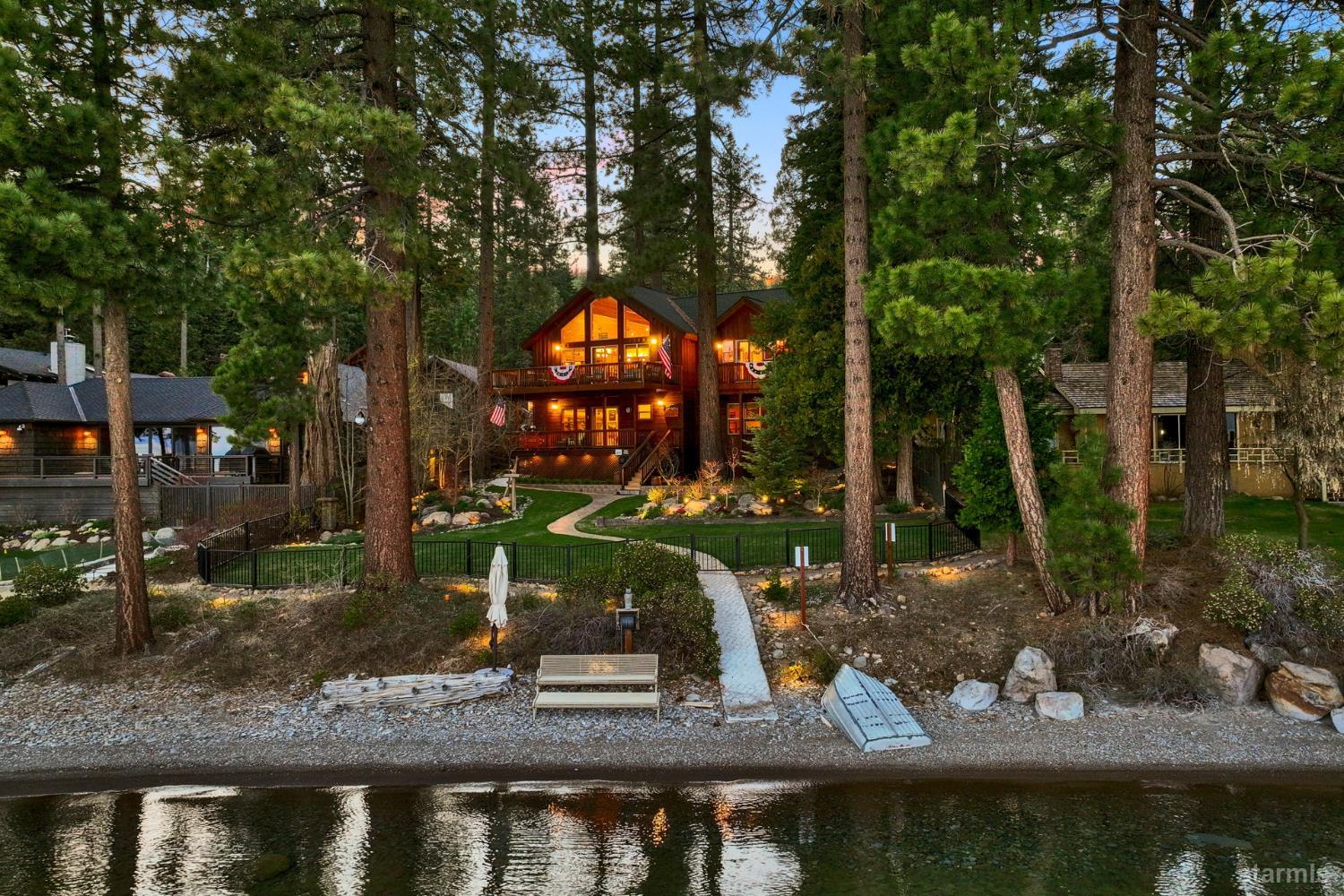 Wooden lakefront home glowing at dusk among pines, with a landscaped path leading to the water.
