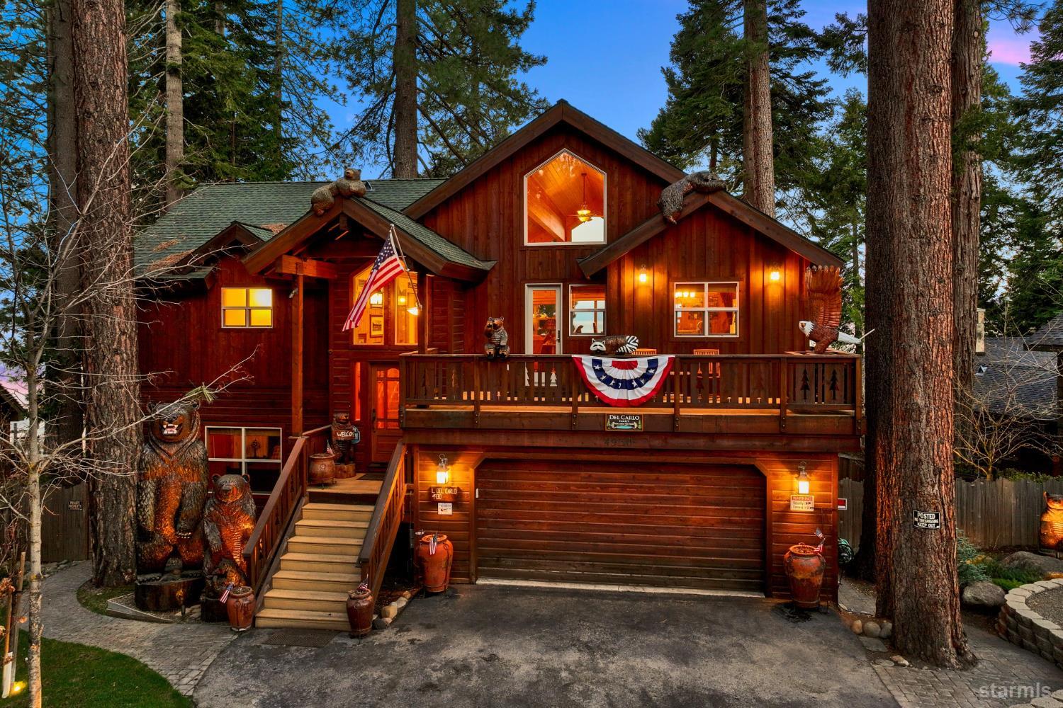 Rustic wood cabin among tall pines, with a lit garage, broad deck, American flag, and carved bear statues.