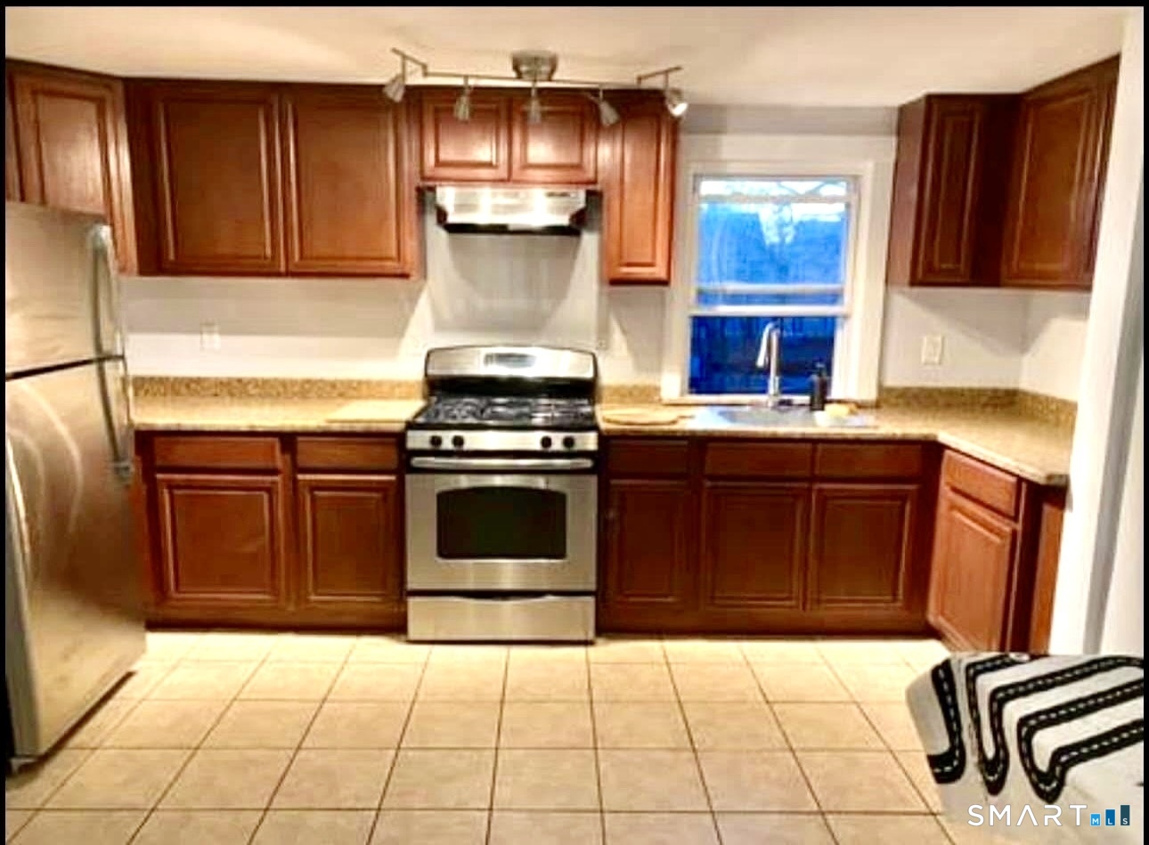 Kitchen with reddish-brown cabinets, stainless-steel appliances, tiled floor, and a window above the sink.