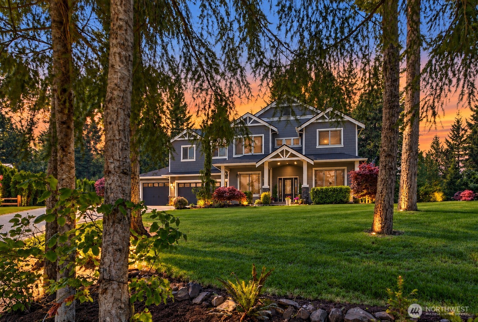 Blue two-story home framed by trees at sunset, with a landscaped lawn and glowing windows.