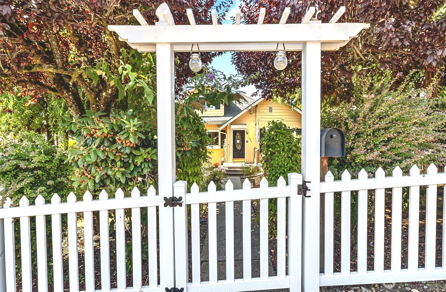 Yellow house behind a white picket gate and pergola, framed by dense landscaping.