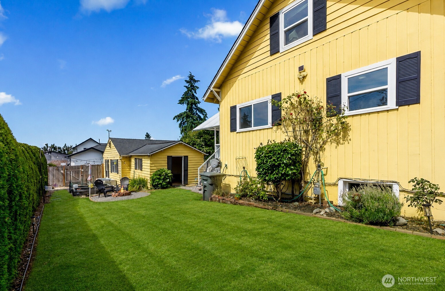 Yellow house beside a broad green backyard, with a small detached outbuilding and patio seating.
