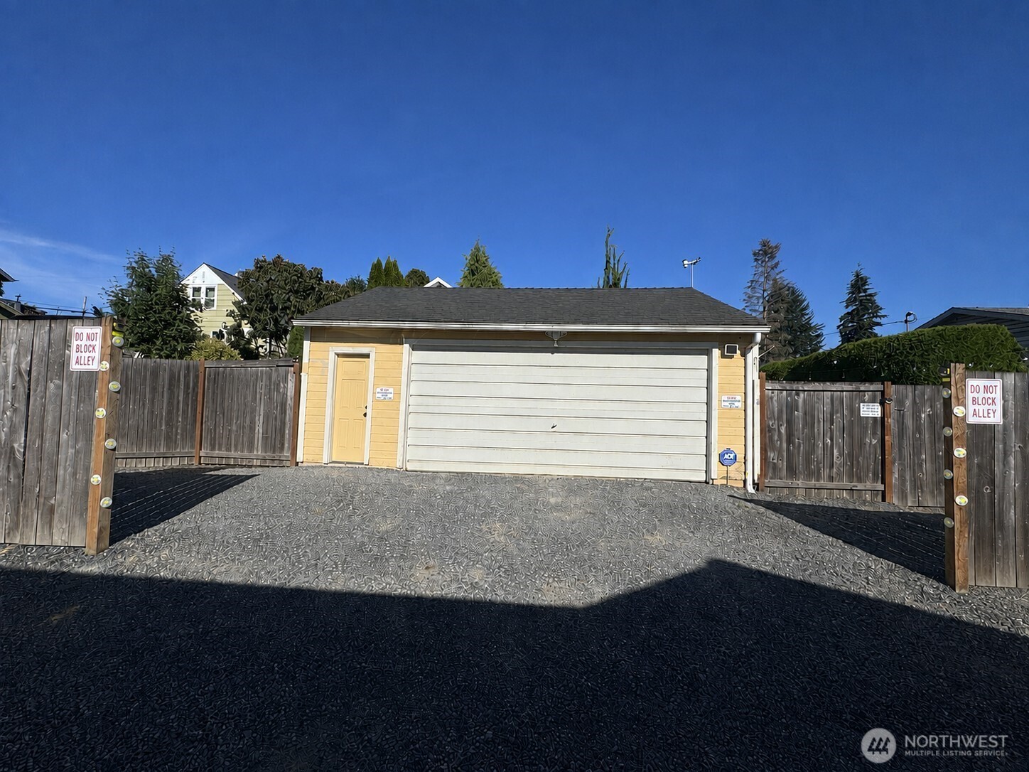 Detached yellow-sided garage with a wide white door, gravel driveway, and wooden alley gates.