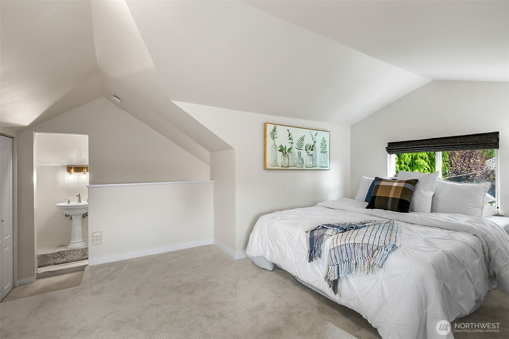 Staged bedroom with a white bed, plaid throw, vaulted ceiling, and bathroom pedestal sink.