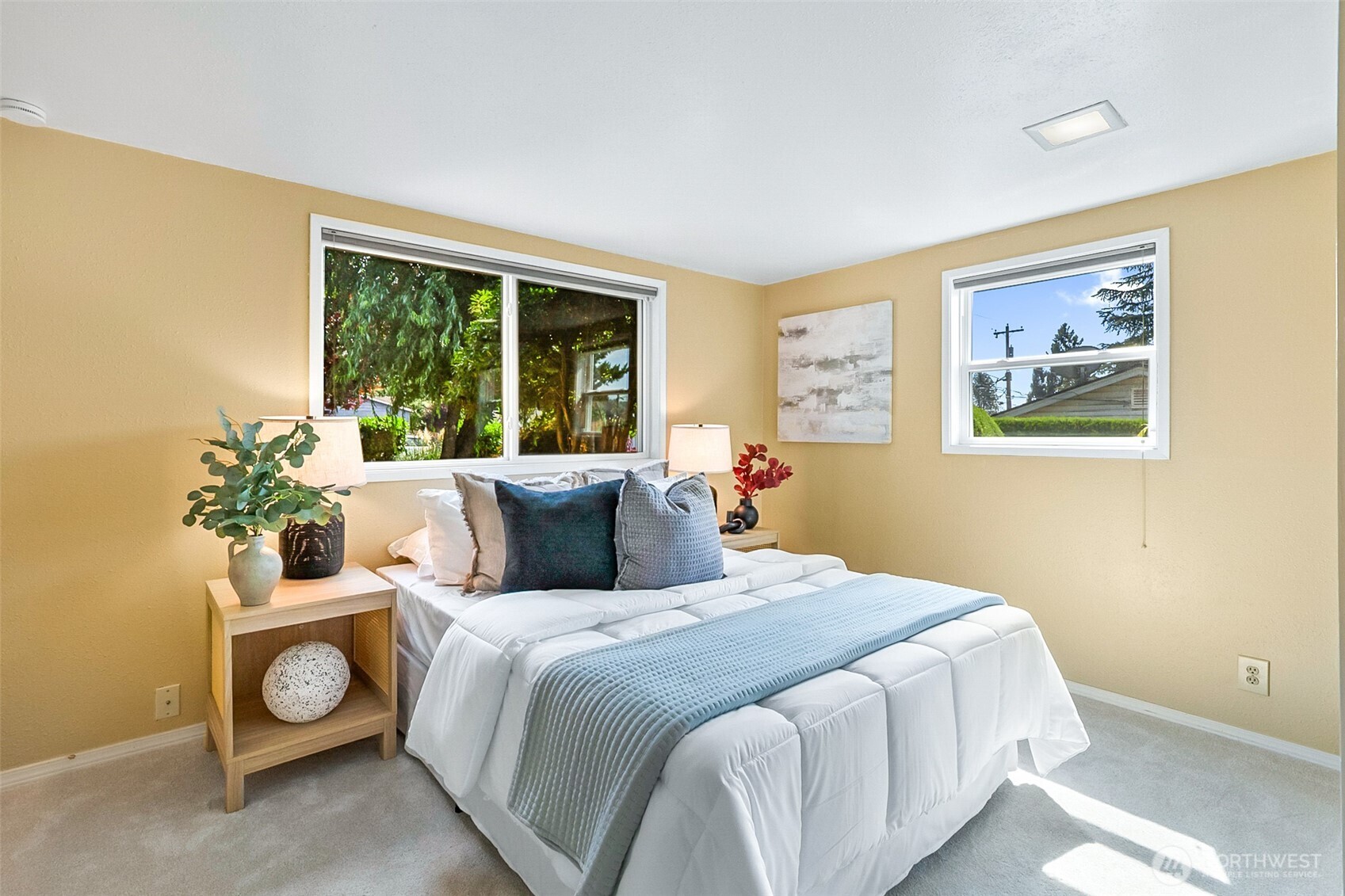 Staged bedroom with a white bed, blue accents, beige walls, and windows overlooking greenery.