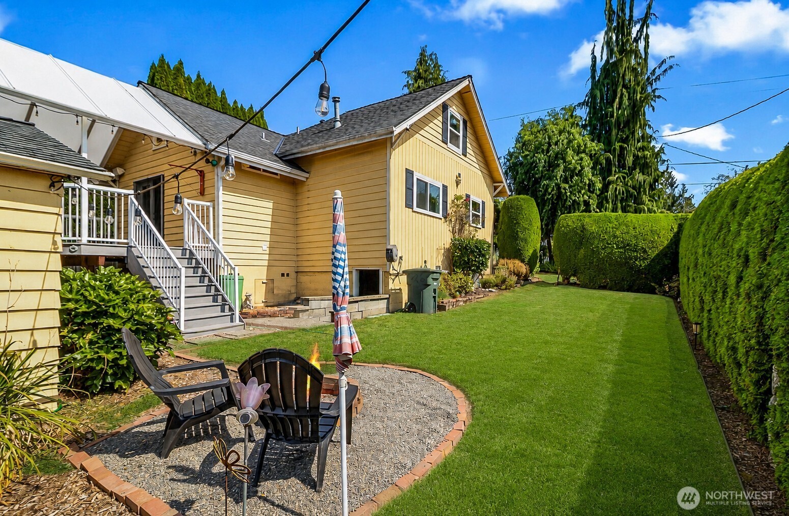 Yellow house beside a green backyard lawn, tall hedges, and a gravel patio with outdoor chairs.
