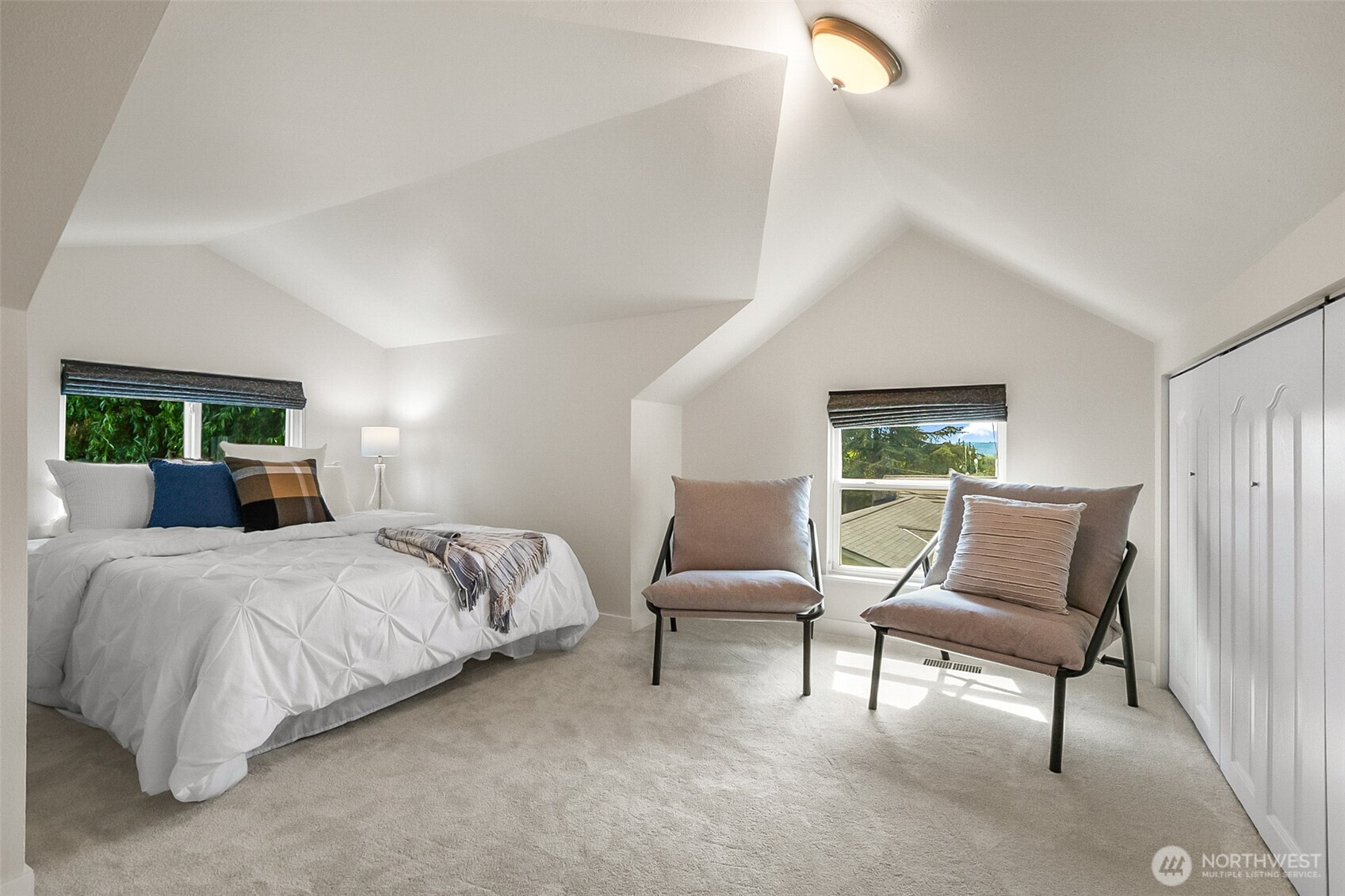 Staged bedroom with a vaulted ceiling, white bed, and two chairs beside a window.