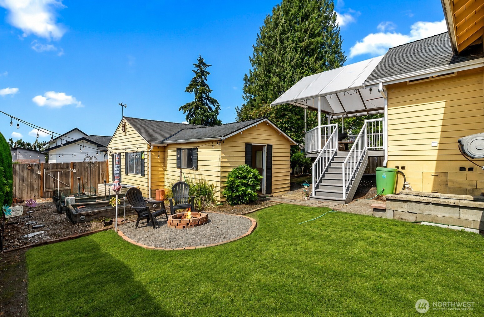 Backyard with a covered raised deck, yellow outbuildings, lawn, and gravel fire-pit seating area.